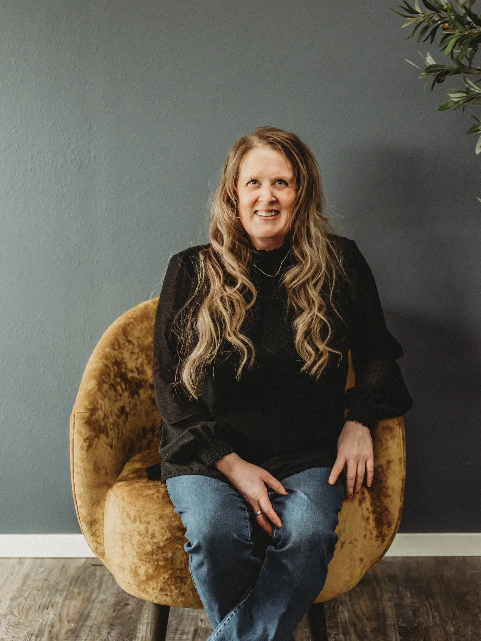 Woman with long, wavy hair sitting on a mustard-colored velvet chair, smiling against a gray wall with part of a green plant visible on the right.