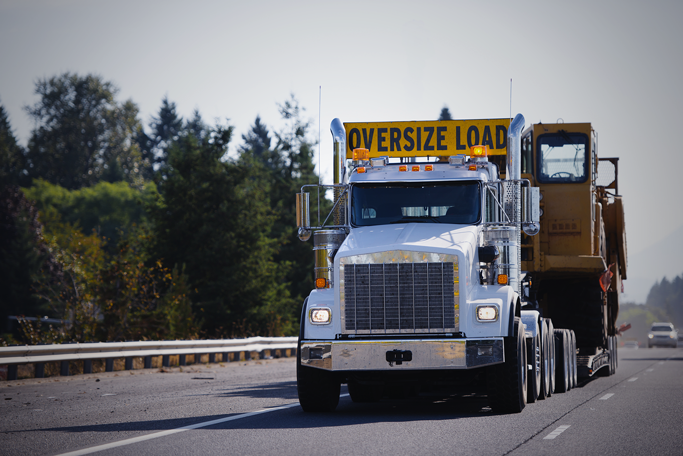 Large white semi-truck with an oversized load sign on top, driving on a highway with trees in the background.