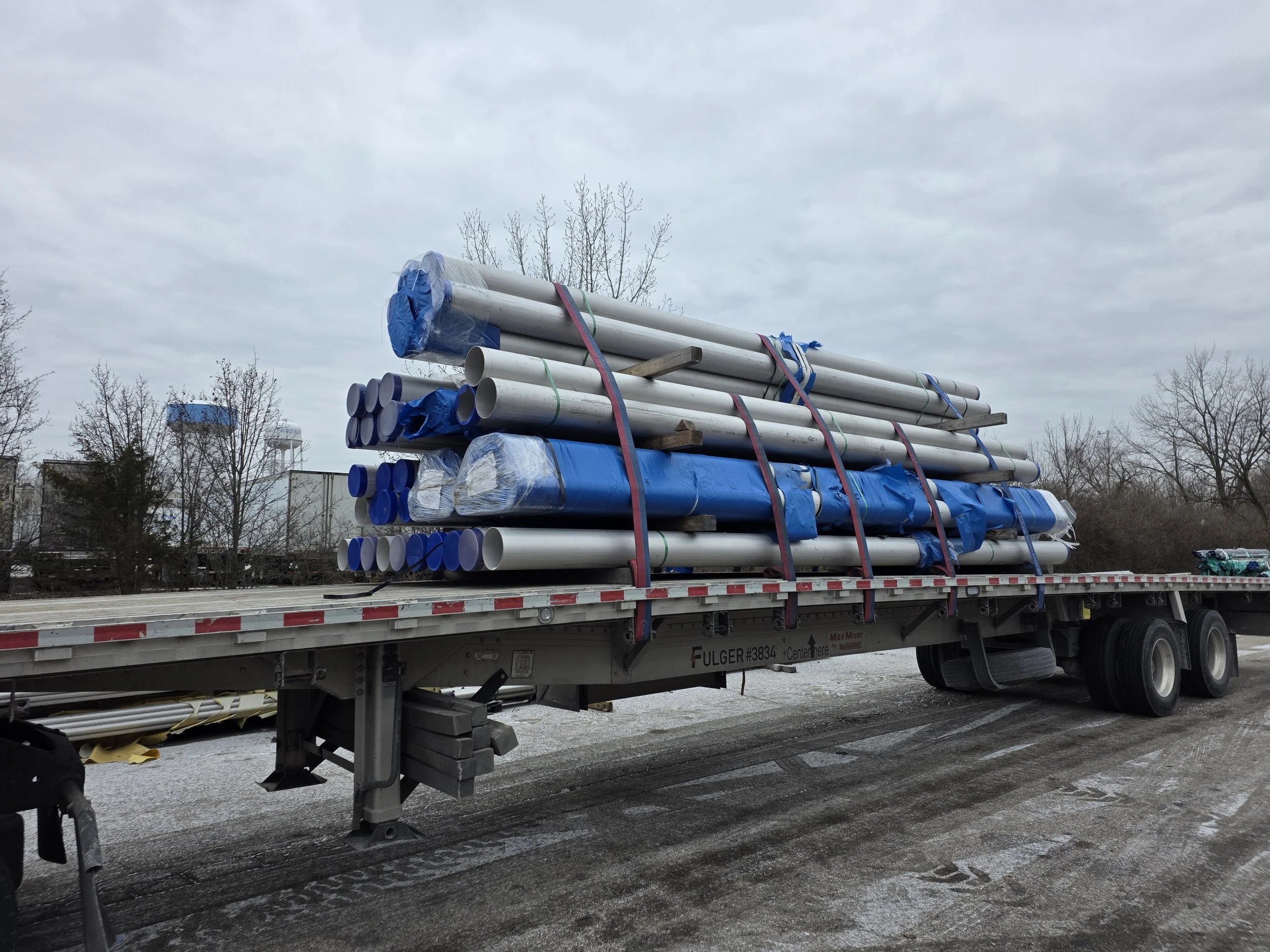 Flatbed truck carrying long metal pipes and wrapped materials, secured with straps, on a cloudy day.