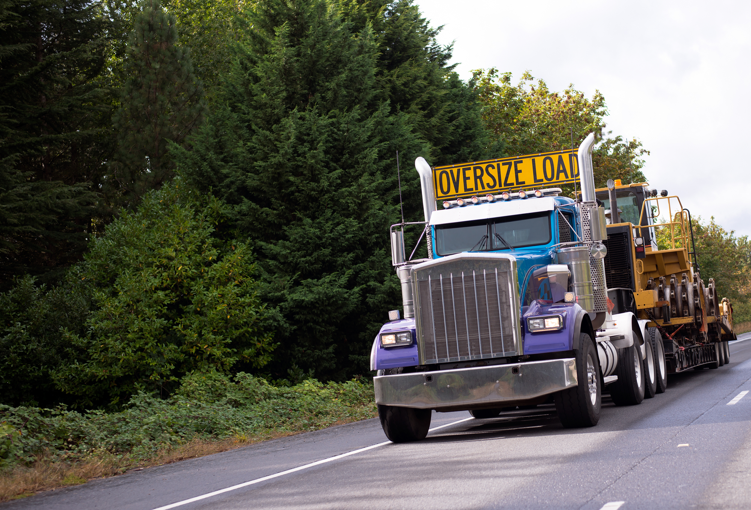 A large semi-truck with a sign saying 'Oversize Load' attached to the front of the truck driving on a highway with trees on both sides.