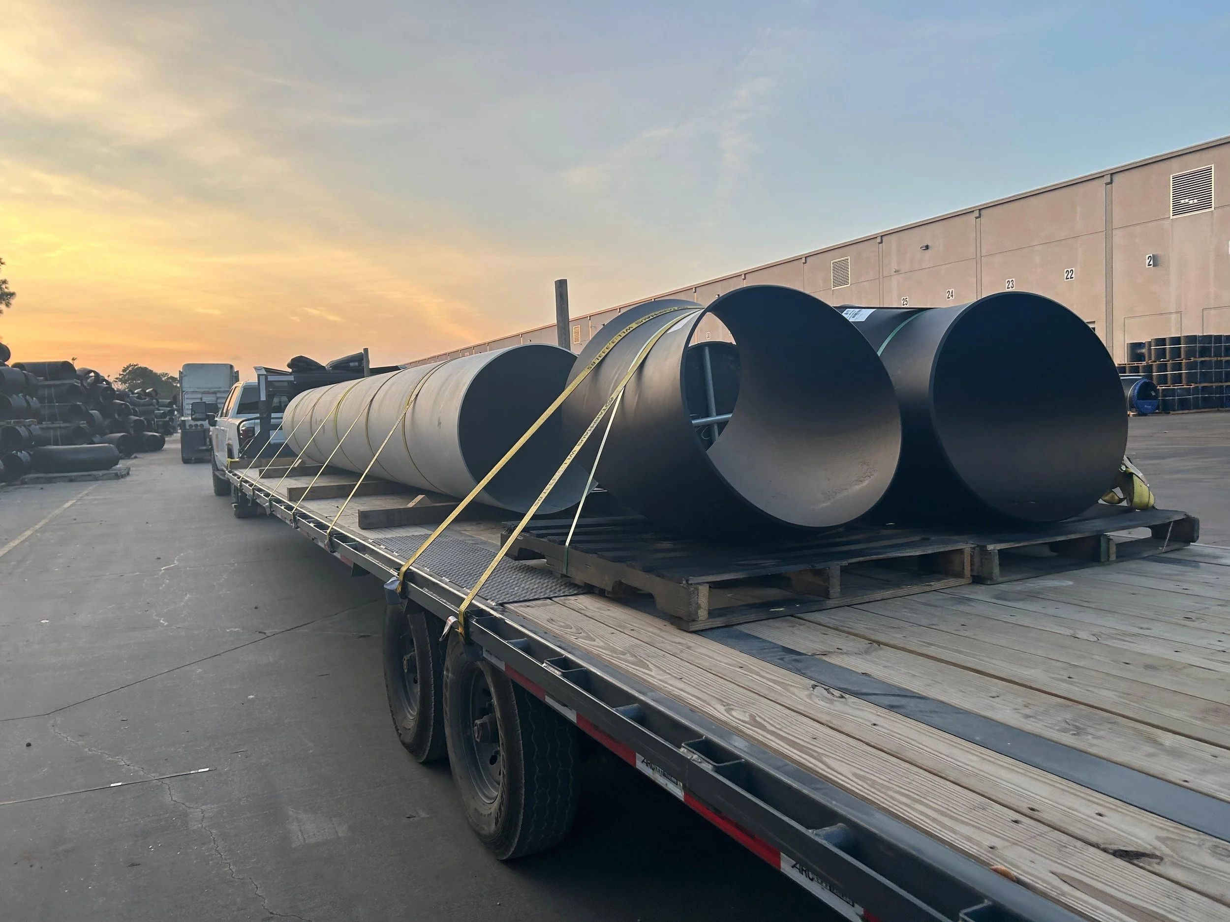 Large black and gray pipes on a flatbed truck at sunset in an industrial area.
