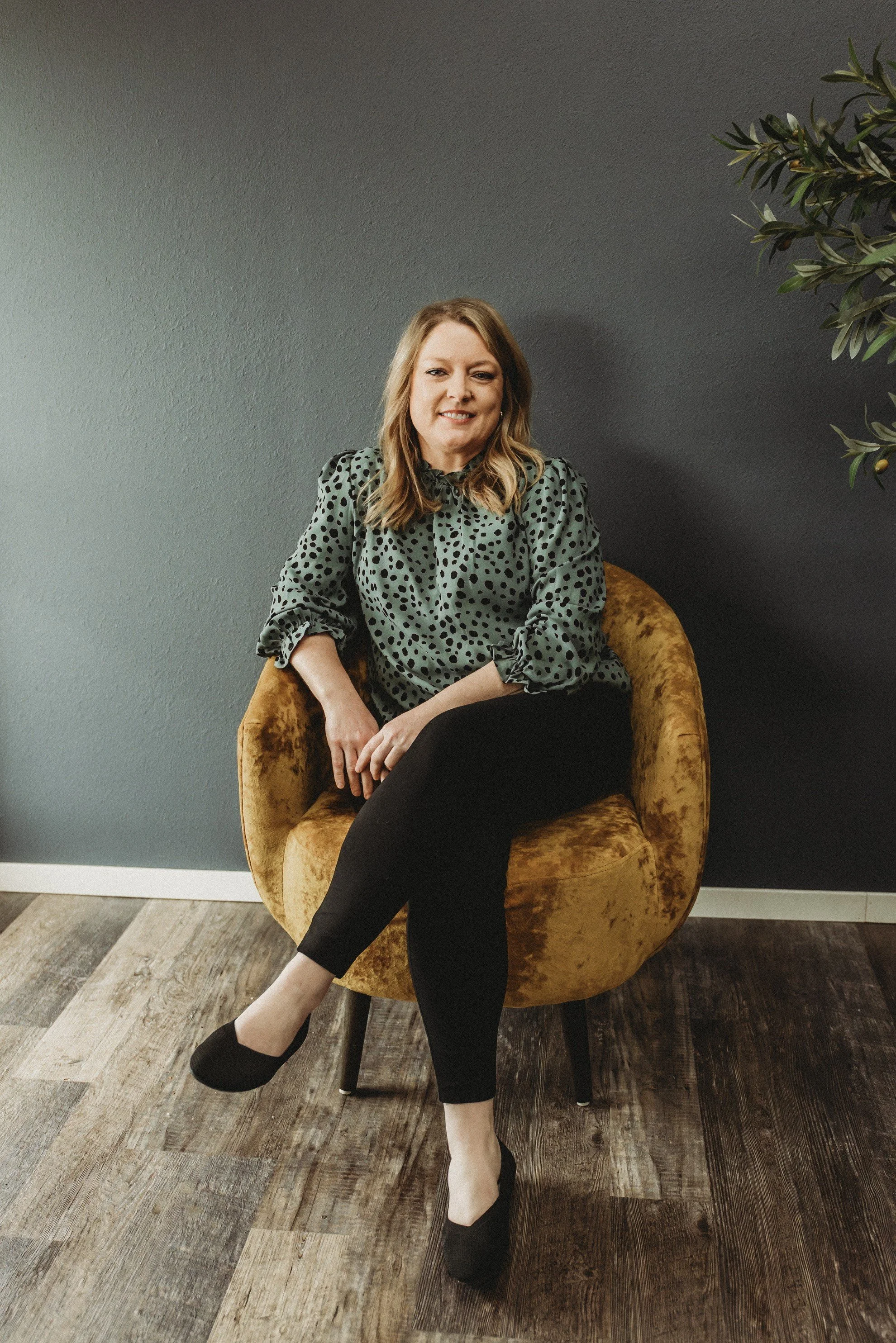A woman with shoulder-length blonde hair sits on a yellow velvet chair, wearing a green polka-dot blouse and black pants, against a gray wall and wooden floor.