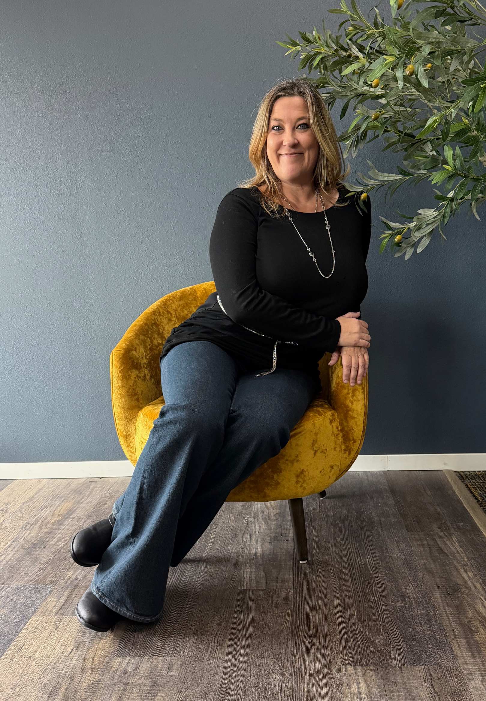 A woman sitting on a yellow velvet chair near a wall with a green plant beside her.