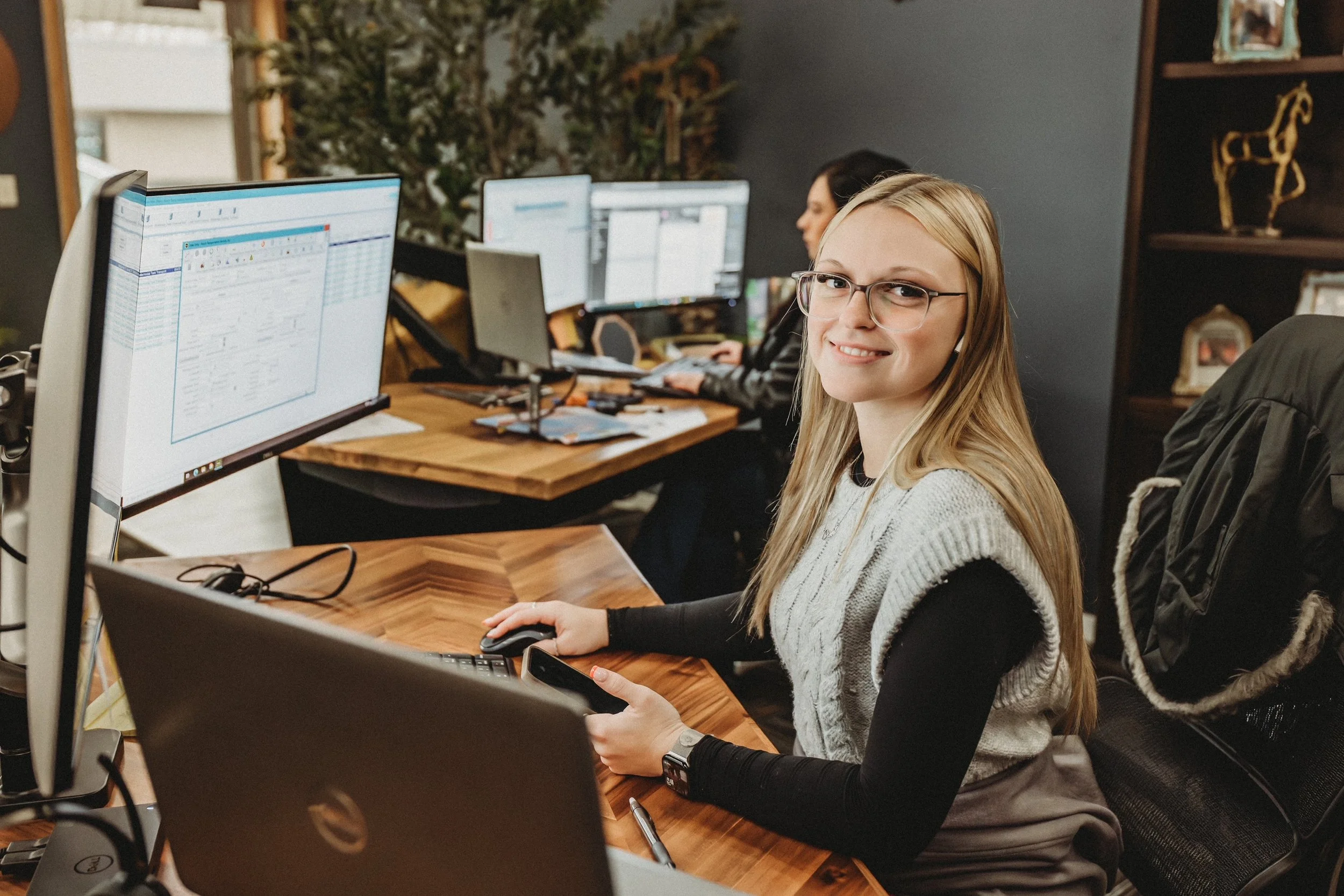 A woman with glasses sitting at a desk with multiple computer monitors in an office setting, smiling at the camera.