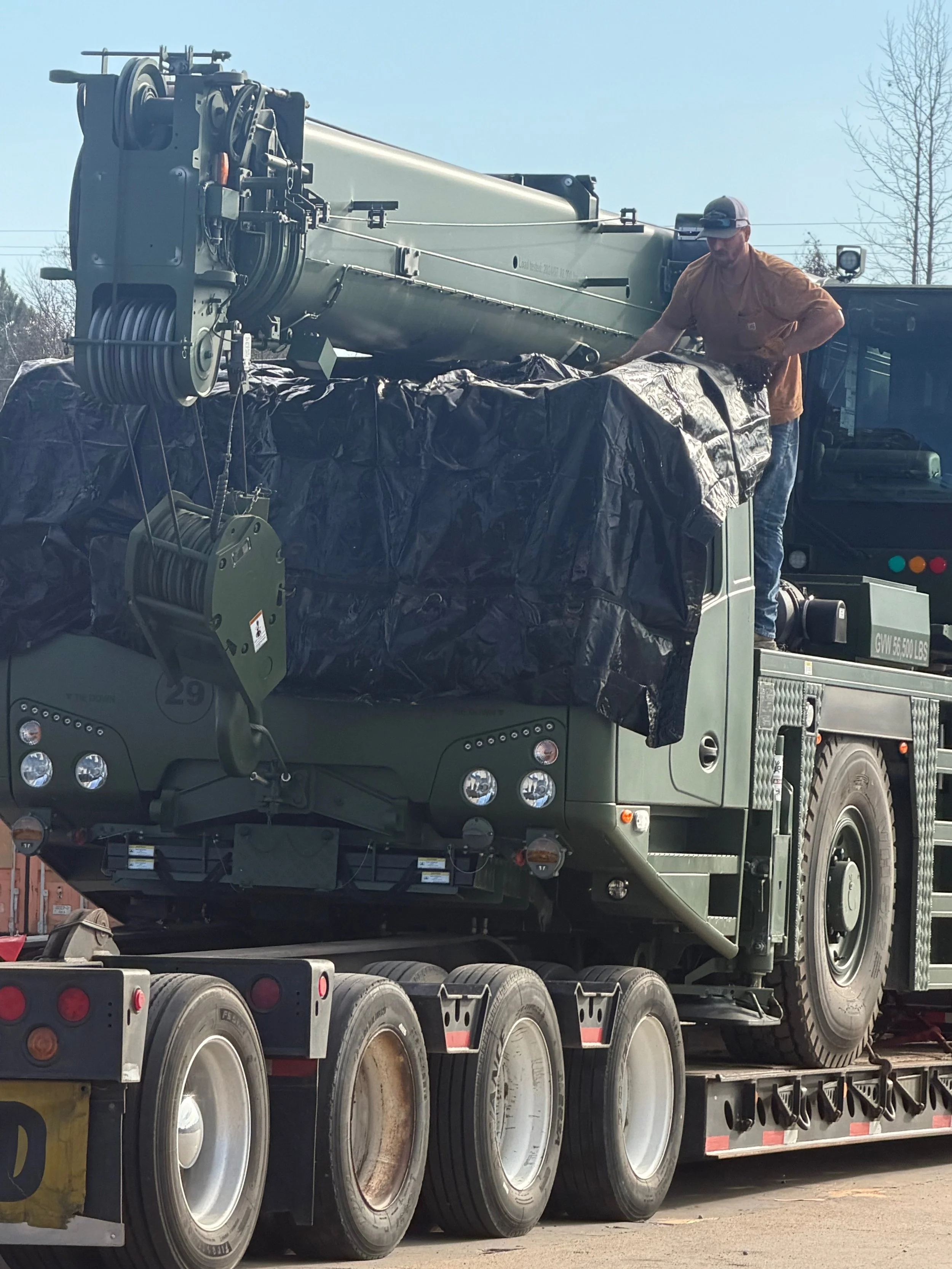 Military missile launcher being transported on a flatbed truck with a man standing on top, securing it. Clear sky, trees in the background.