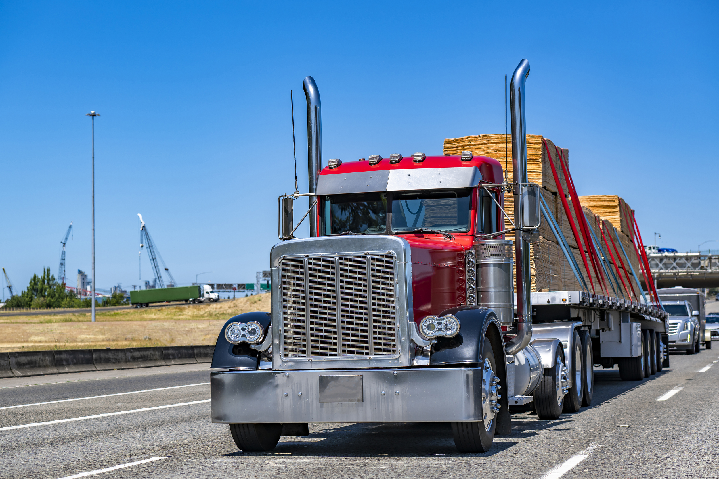 A red semi-truck carrying stacked wooden pallets on a highway under a clear blue sky.