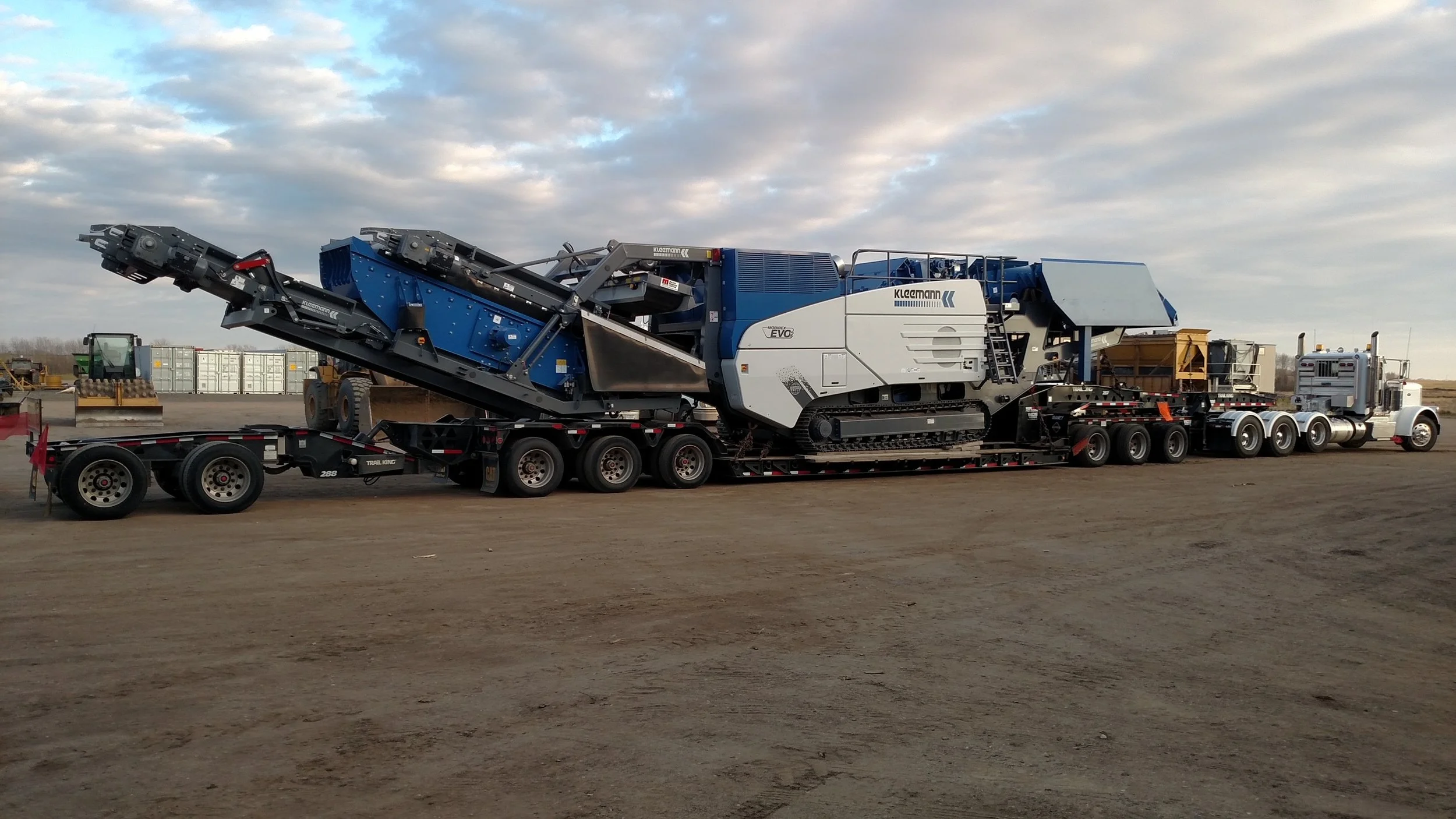Large construction machine on a flatbed trailer, with a blue and gray body, situated on a dirt lot under a partly cloudy sky.