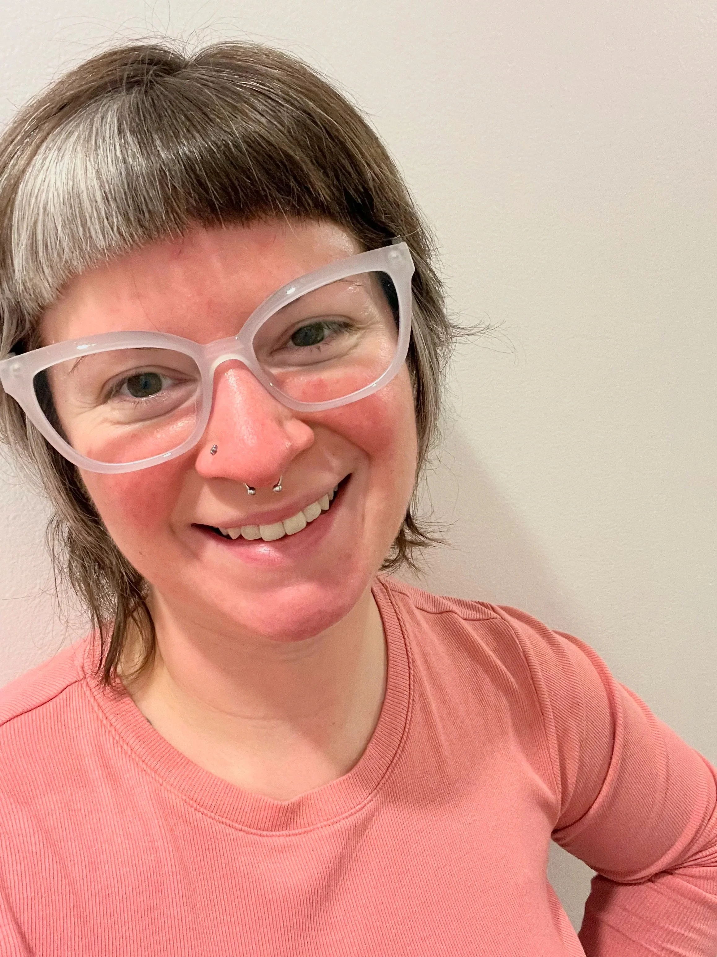 A smiling woman with short, tousled brown hair, wearing clear glasses and a nose ring, dressed in a pink shirt, standing against a plain light-colored wall.