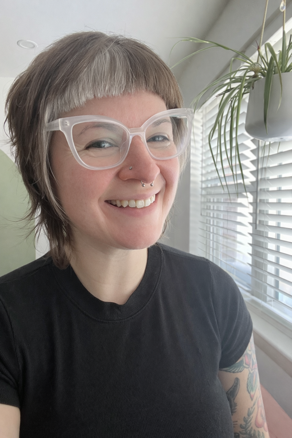 A woman smiling indoors near a window with horizontal blinds and a hanging plant.