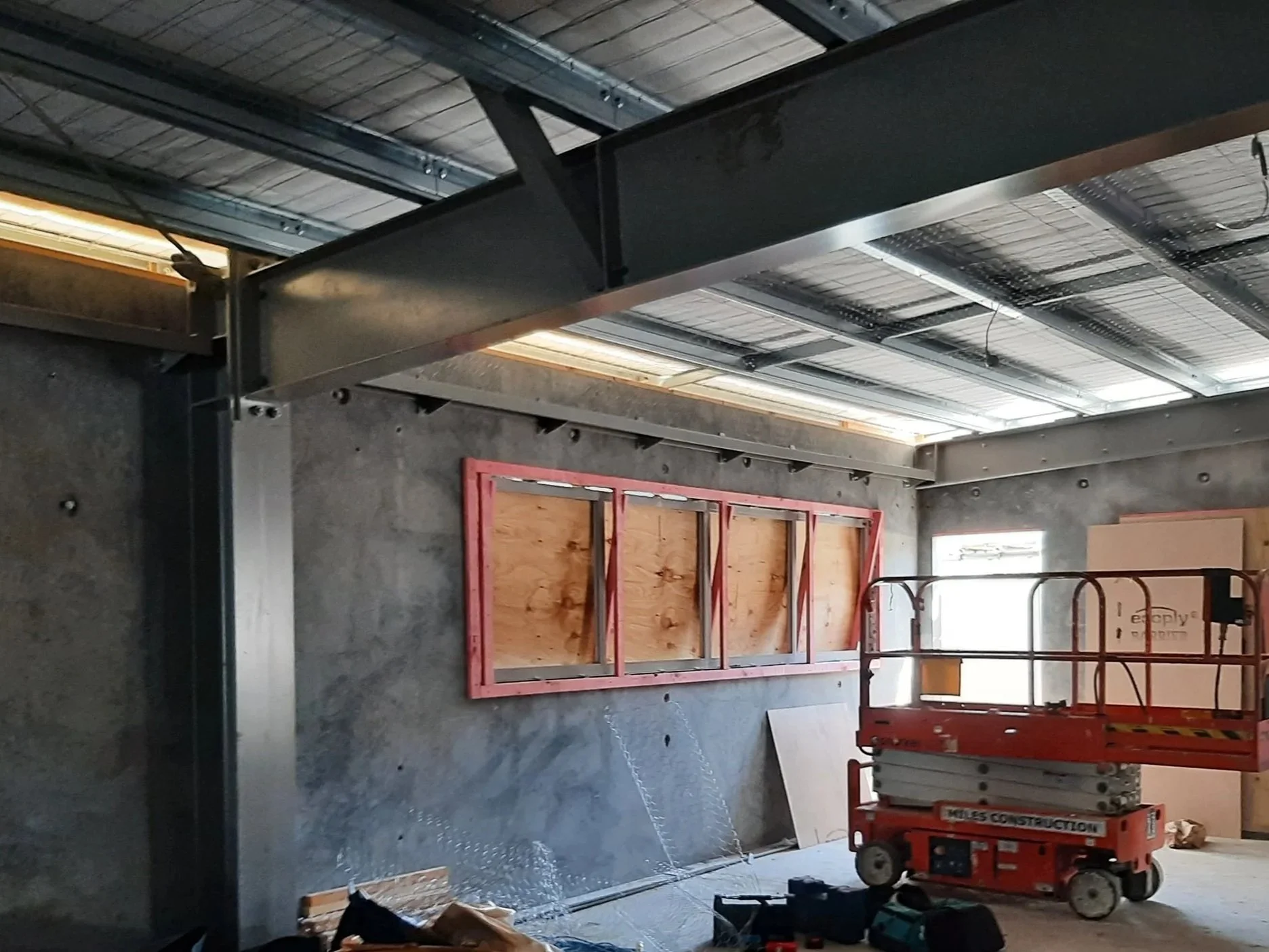 Construction site with a concrete wall, a window frame attached to the wall, and a scissor lift in the room. The ceiling has metal roofing and skylights, and there are construction tools and materials scattered on the floor.