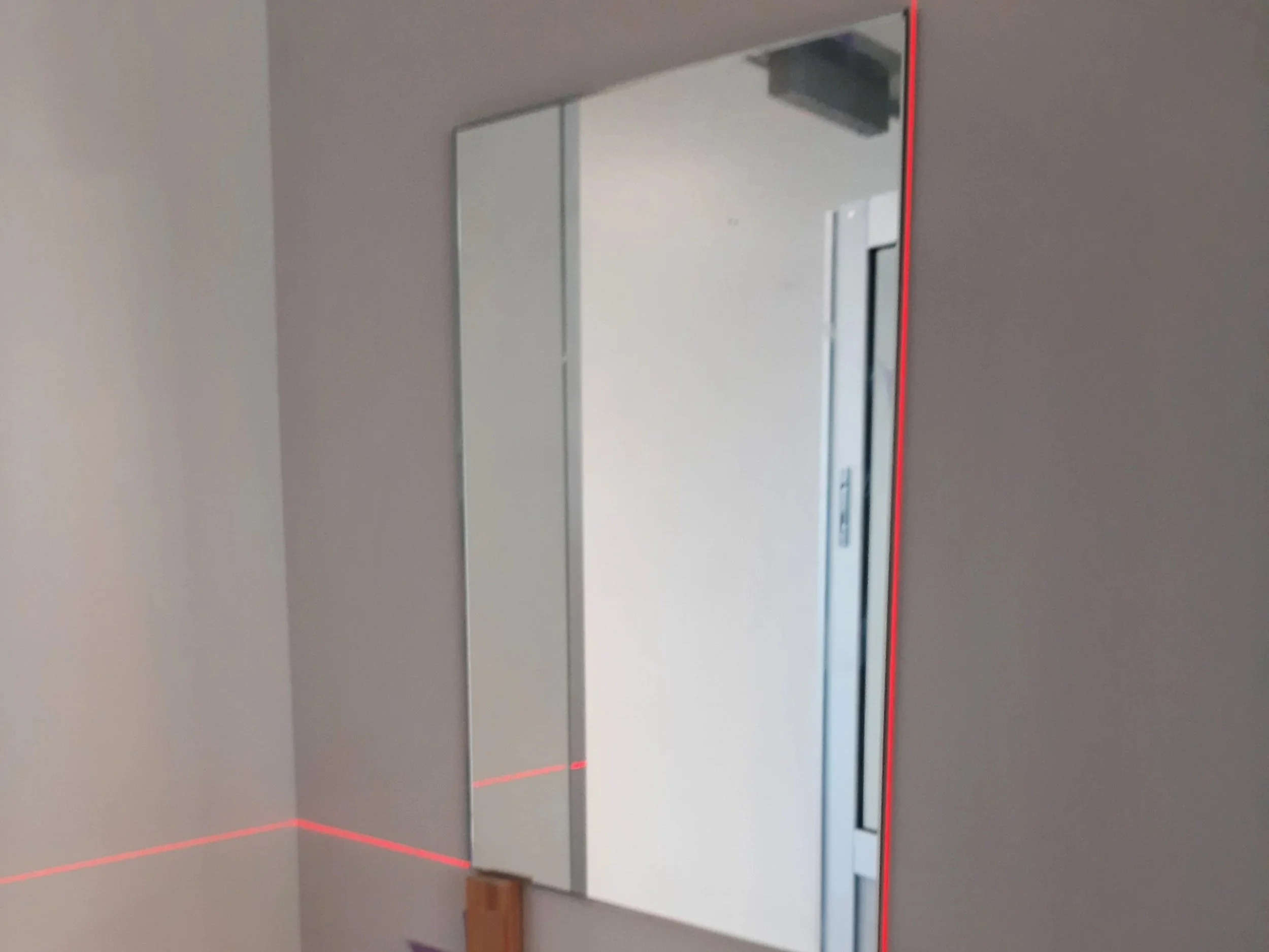 A mirror with a red laser line projected across the bottom and right edges, mounted on a gray wall near a door.