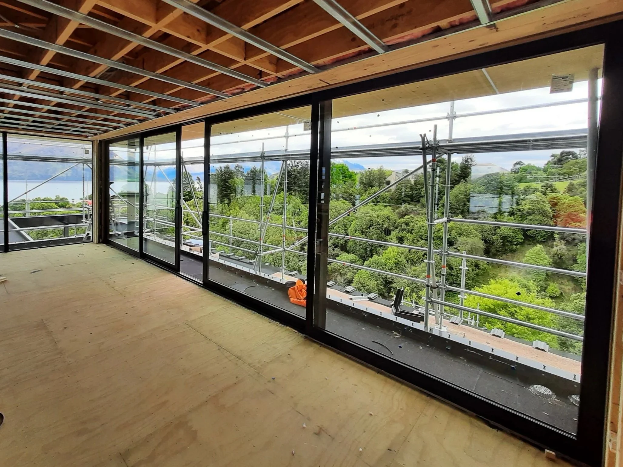 Interior of a building under construction with large glass windows and frames outside, overlooking a green landscape and distant view.