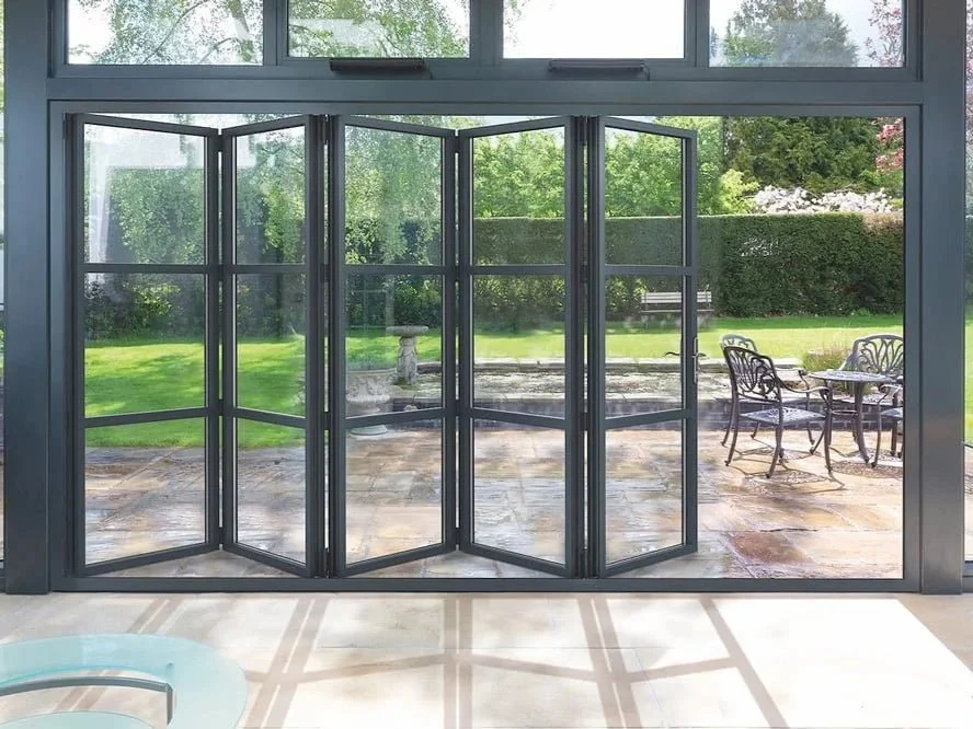 Foldable glass doors leading to a patio with outdoor table and chairs, overlooking a garden with green grass and trees.
