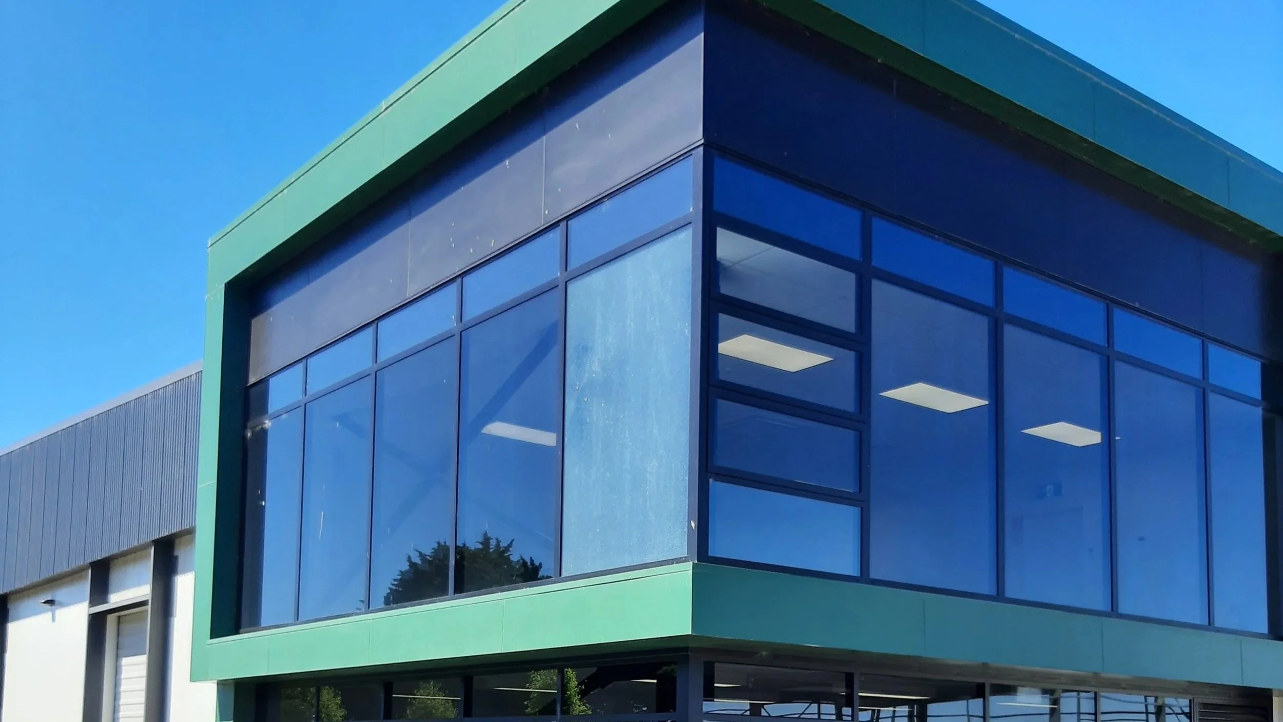 Modern commercial building with large clear and clean glass windows and green framing, under a clear blue sky.