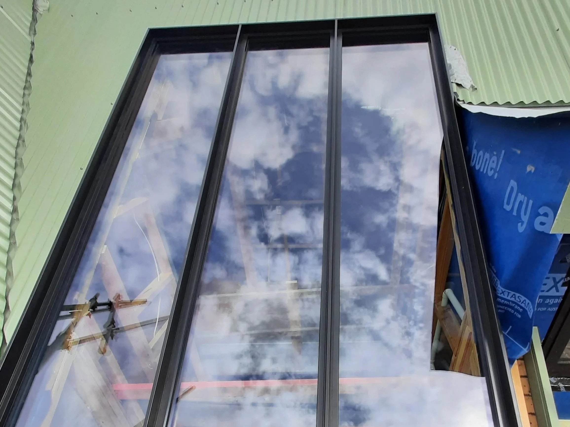 Reflective glass window with sky and clouds reflecting on it, framed by black metal, with some construction materials visible around it.
