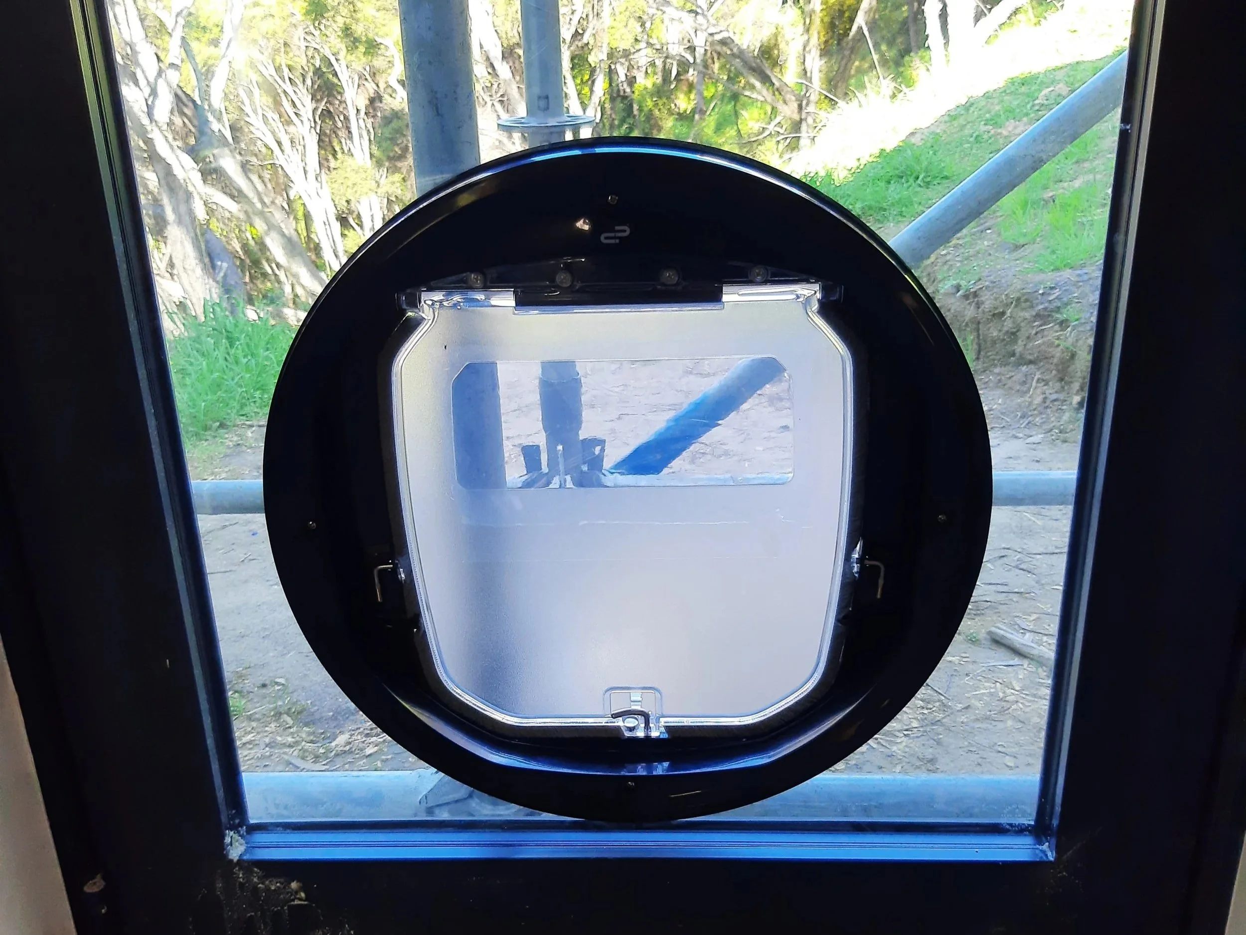 A circular pet door with a transparent flap installed in a glass door, looking out to trees and greenery outside.