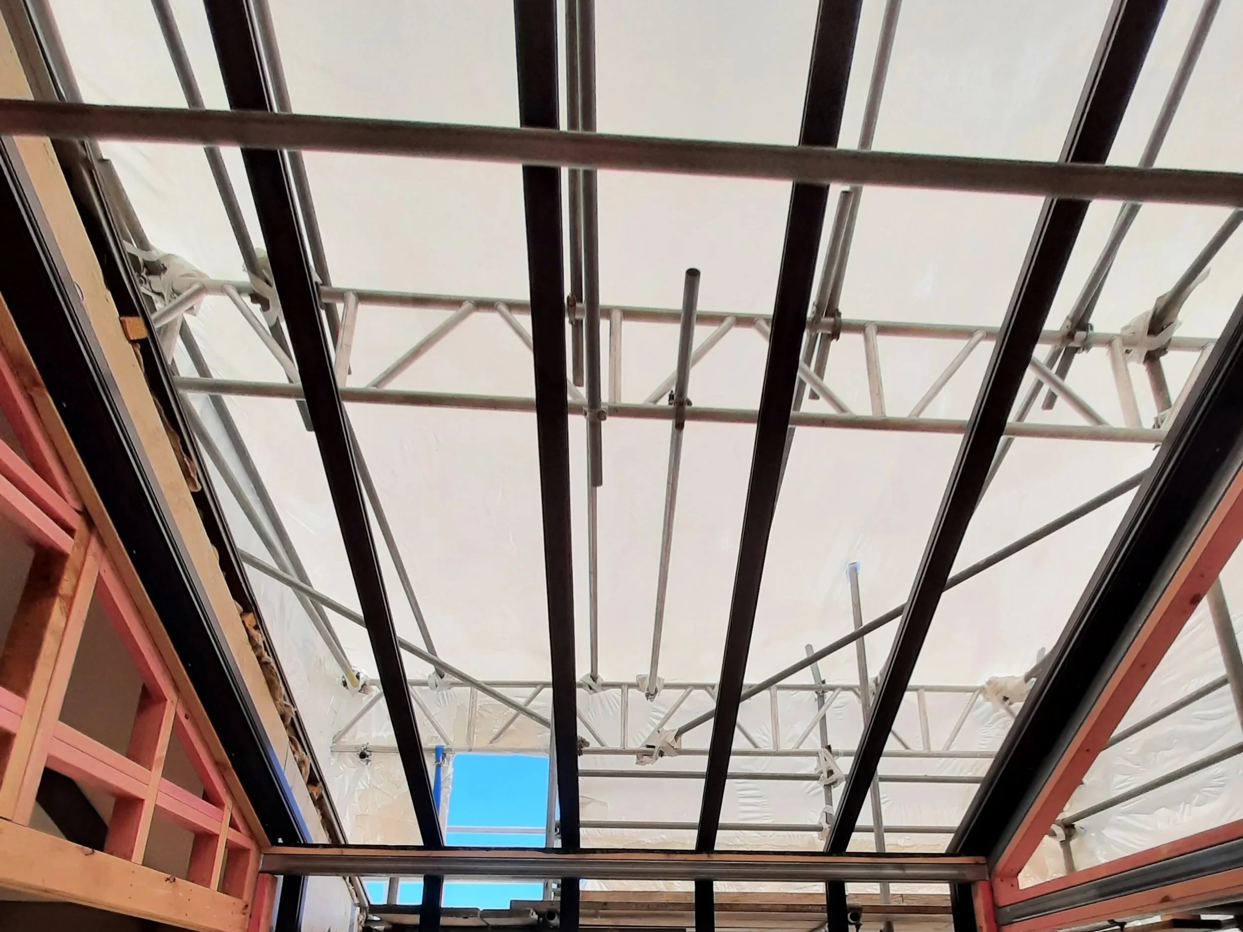 Construction site with scaffolding and framework seen from below, with a window opening showing blue sky.