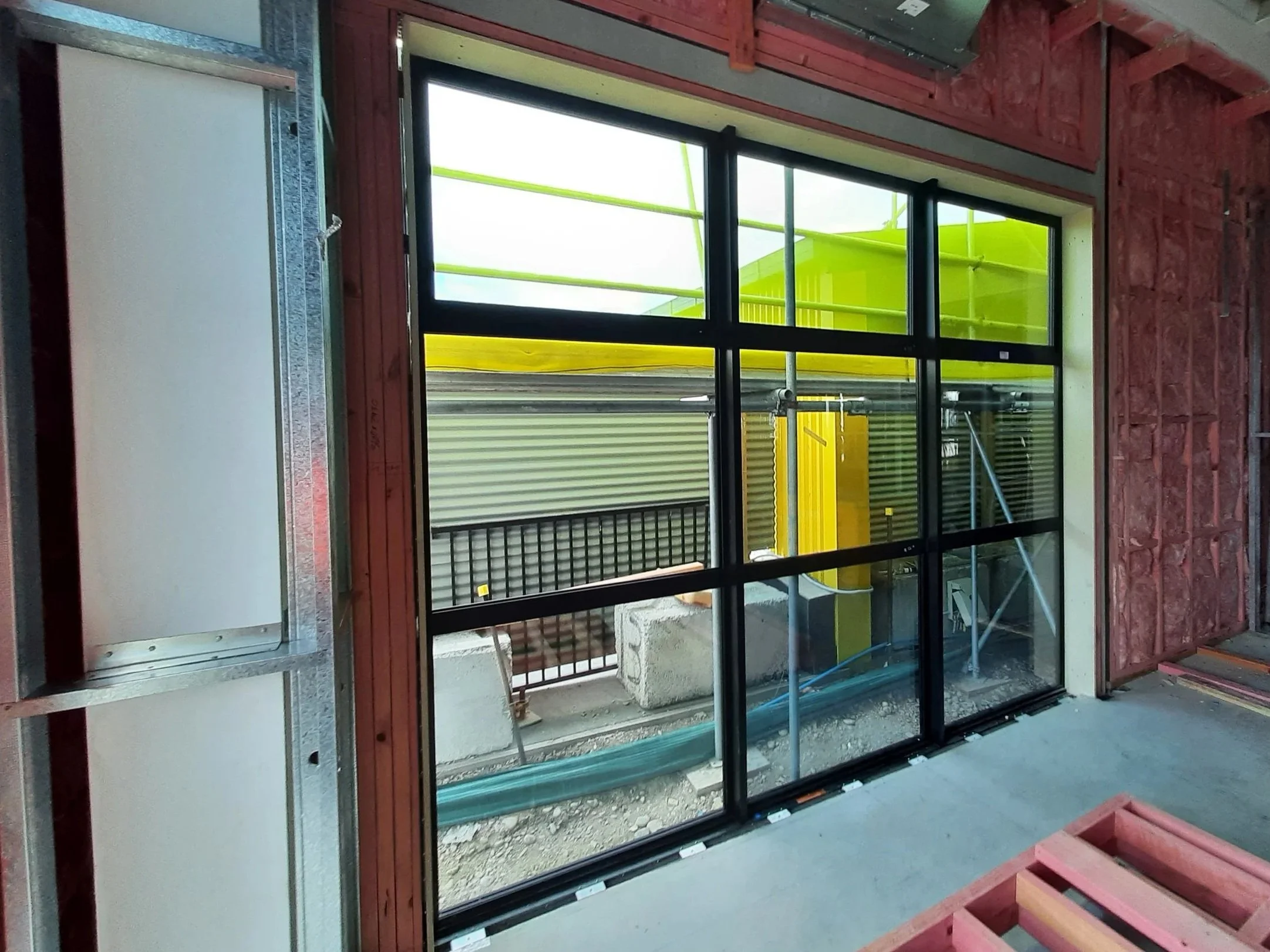 Interior view of a building under construction, showing a large window with black frame and glass panes, partially installed. Construction materials, scaffolding, and building supplies are visible outside through the window.