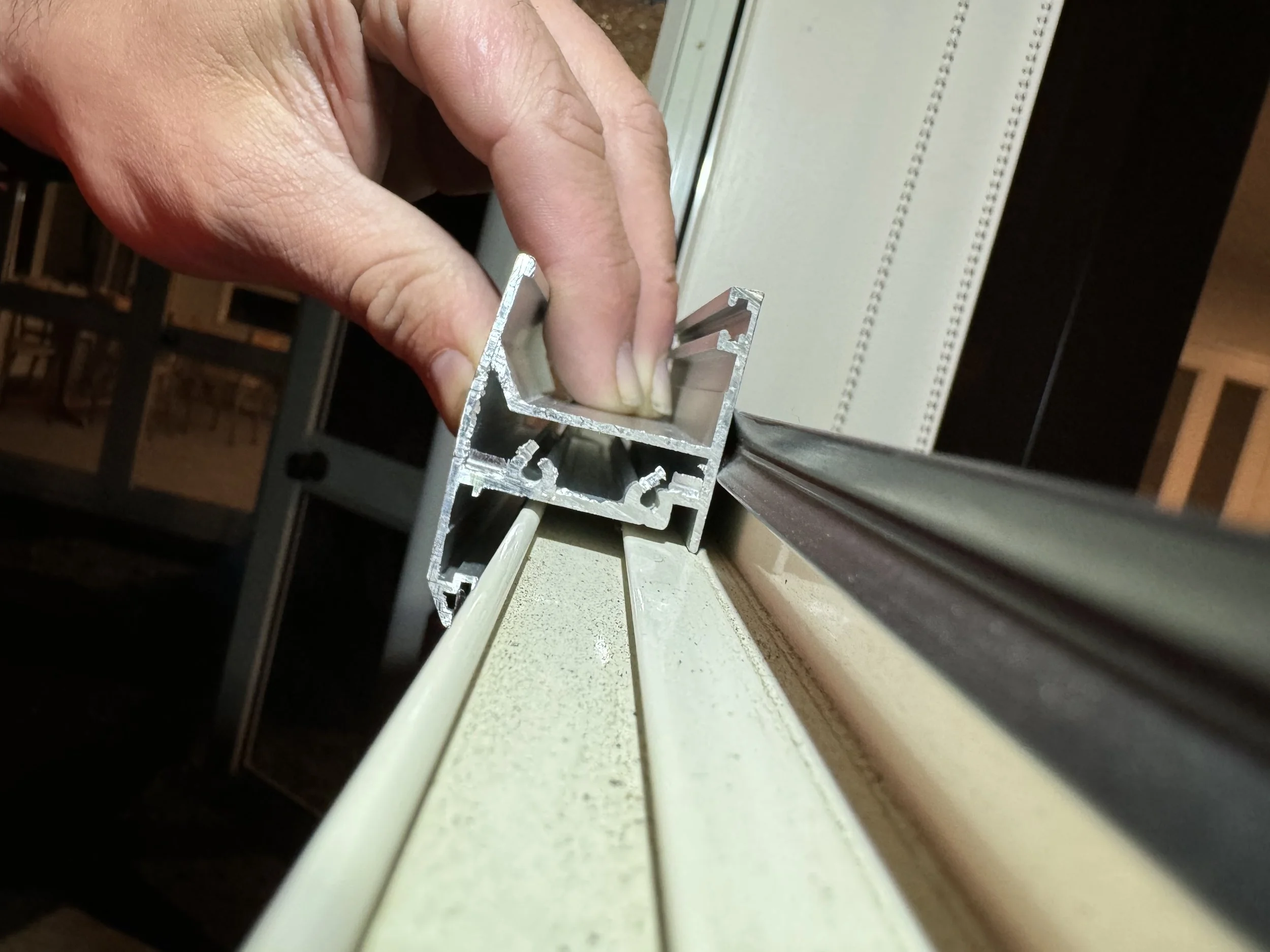 Close-up of a hand holding an aluminium window frame track, positioned along a window sill in a room under retrofit double glazing works.