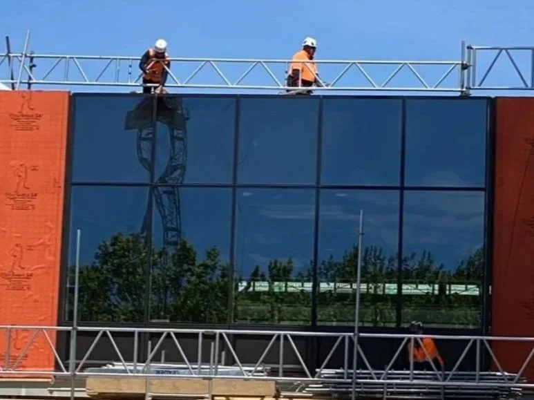 Two construction workers wearing safety helmets working on the roof of a glass building under construction.