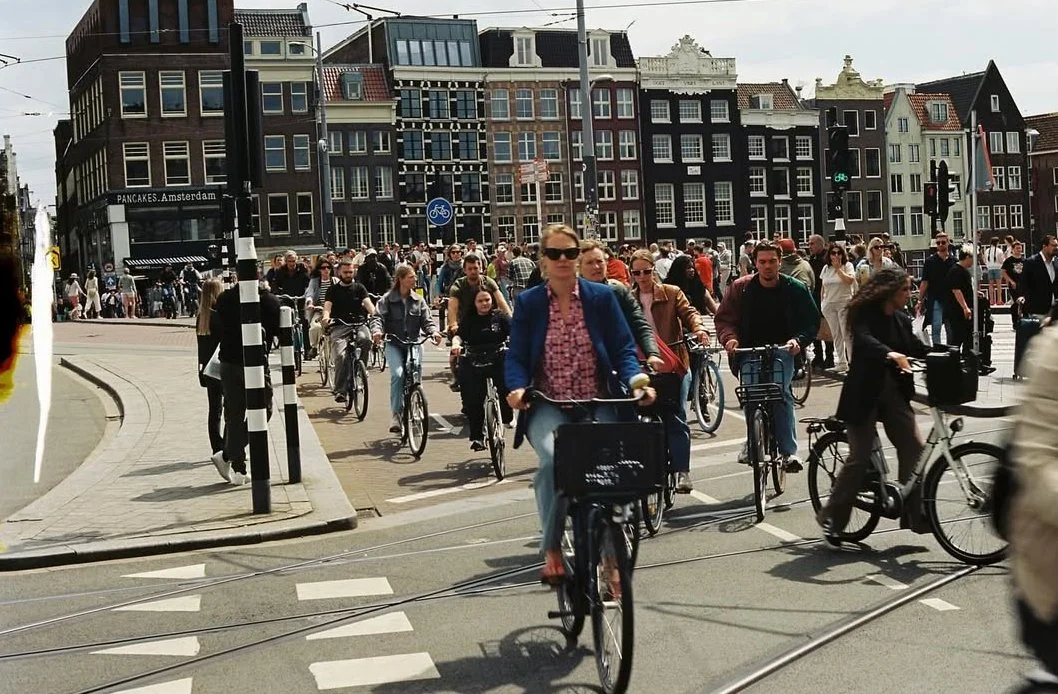 A busy street scene in Amsterdam with many people riding bicycles and walking, in front of traditional Dutch buildings.