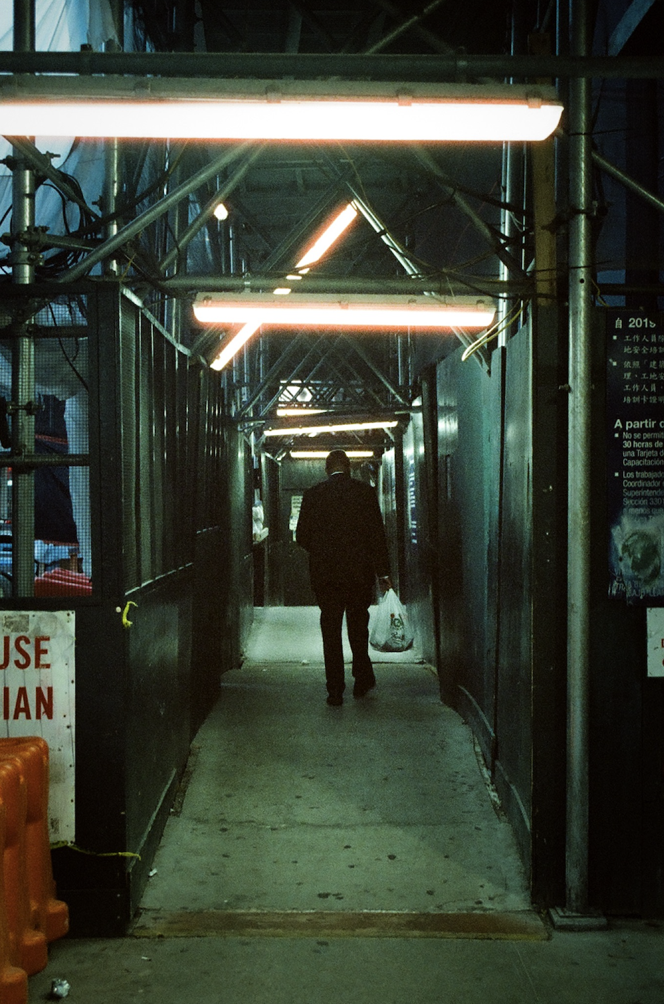 A person walking through a dimly lit construction tunnel at night, carrying a plastic bag, with illuminated fluorescent lights overhead.