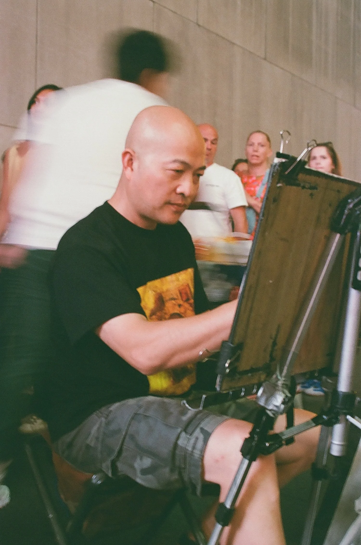 A man drawing or painting on an easel while sitting on a chair, with a group of people standing in the background, in an indoor setting.
