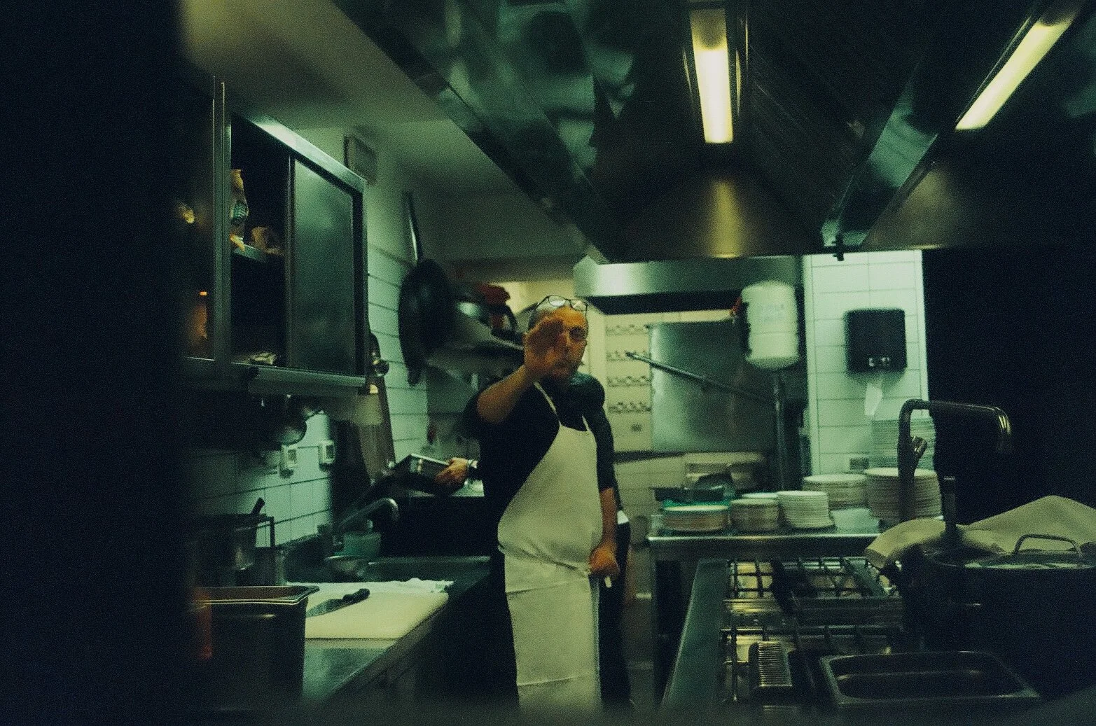 A person in a kitchen with a white apron and glasses on the head, waving or reaching towards the camera. The kitchen has shelves with plates, a stovetop, and various kitchen equipment.