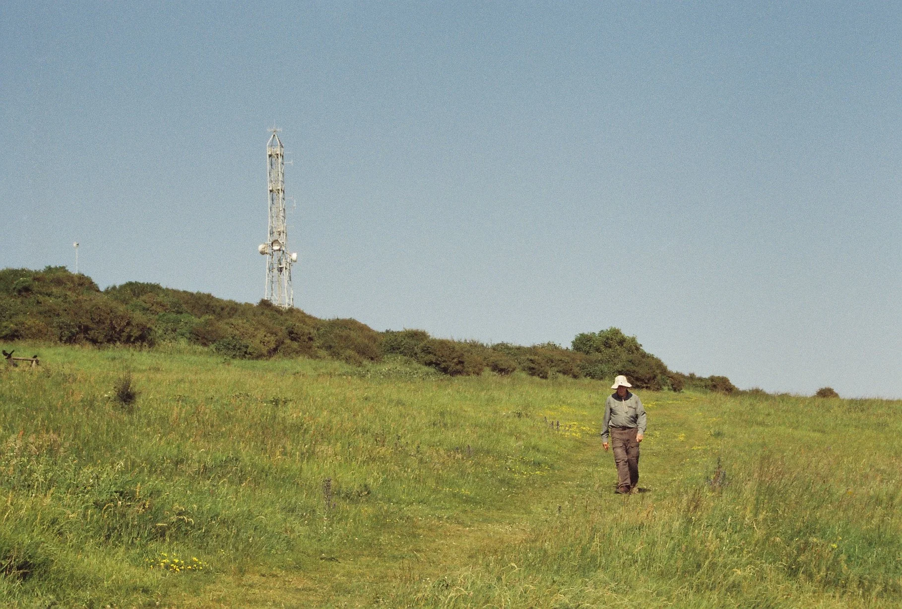 A person walking through grassy field with a hill, radio tower, and blue sky in the background.