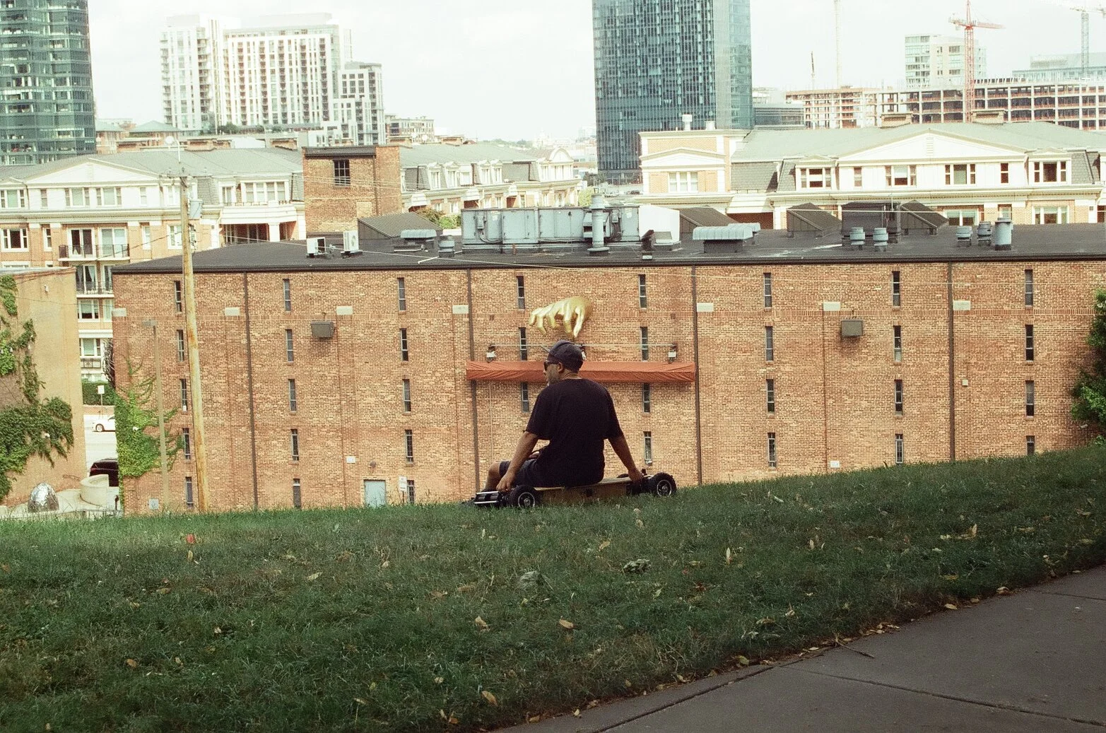 A person sitting on a skateboard on a grassy hill, overlooking a cityscape with various buildings, including a brick building with a white animal statue on the roof.