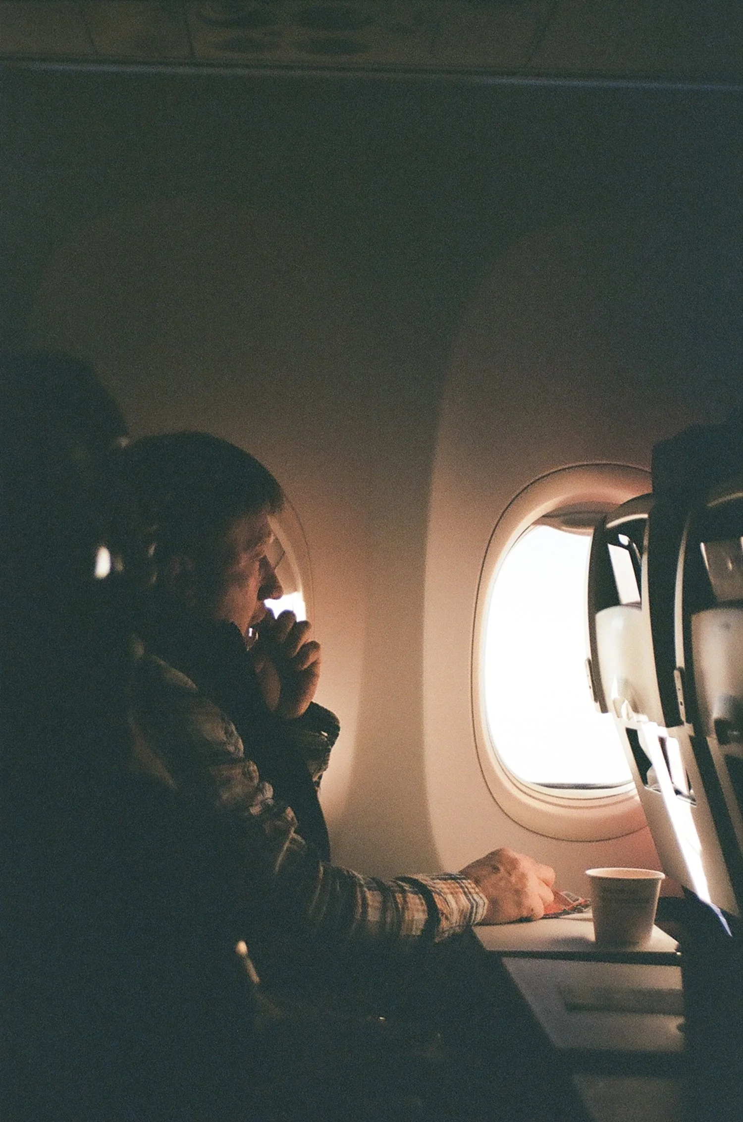 A person sitting by an airplane window, looking out. The person is wearing a jacket and has a book or notebook on the tray table in front. A paper cup is also on the tray table. The scene is dimly lit, with natural light coming from the window.