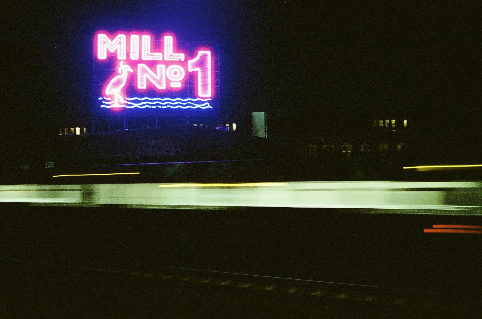 Neon sign reading 'MILL NO 1' with a flamingo and water design, at night with blurred vehicle light trails in the foreground.
