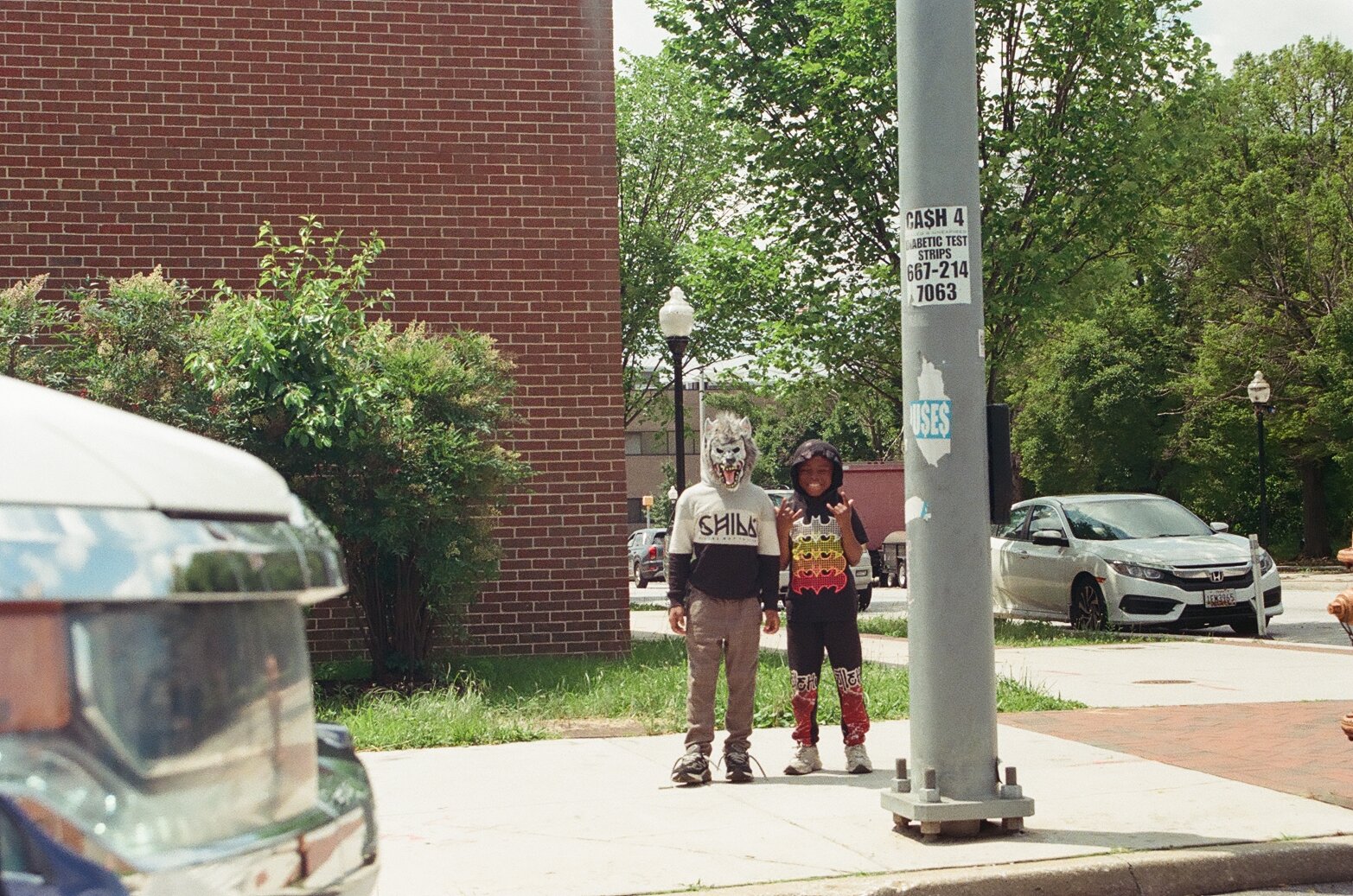Two children standing on a sidewalk in front of a brick building, one wearing a wolf mask and the other smiling, with cars and trees in the background.