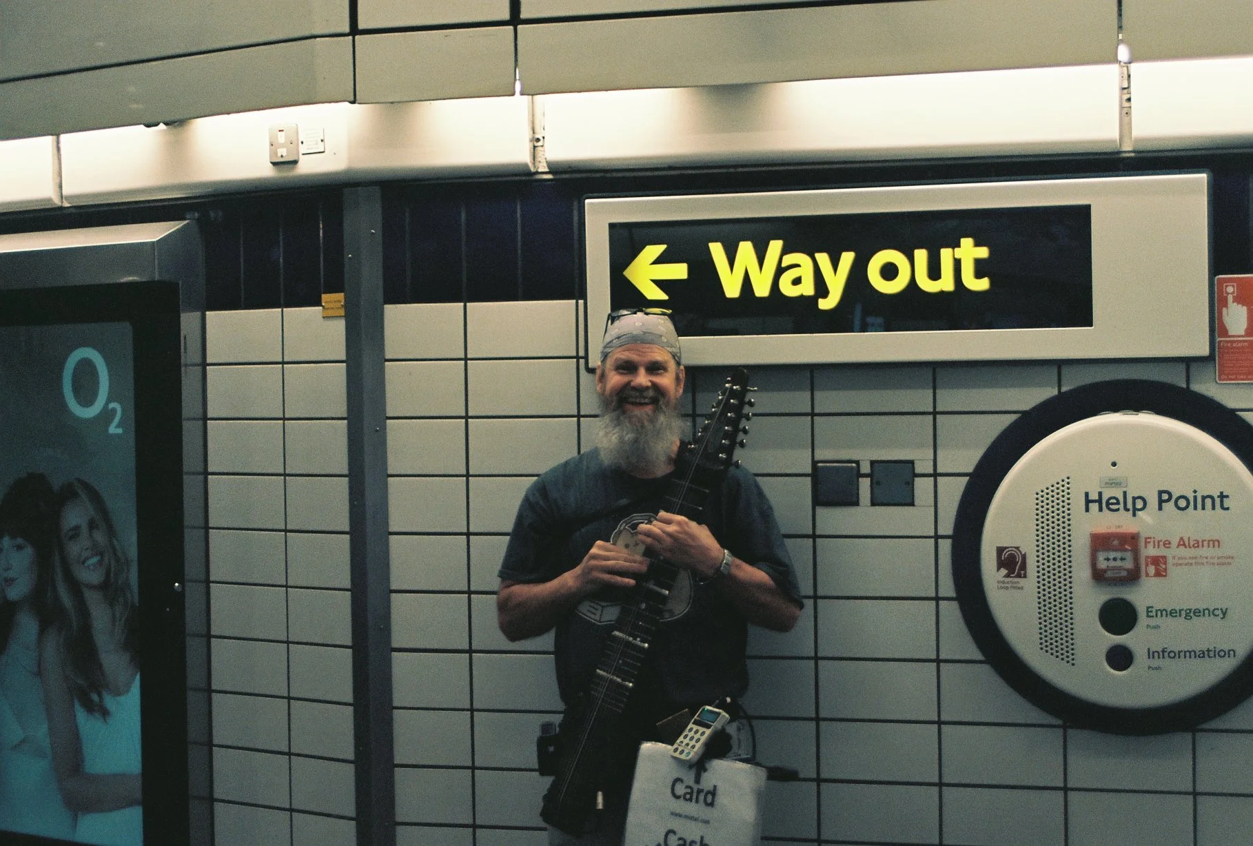 A man with a gray beard, wearing a cap, holding a guitar, smiling at the camera inside a subway or train station with tiled walls. There is a sign pointing left that says 'Way out' and a help point on the wall.