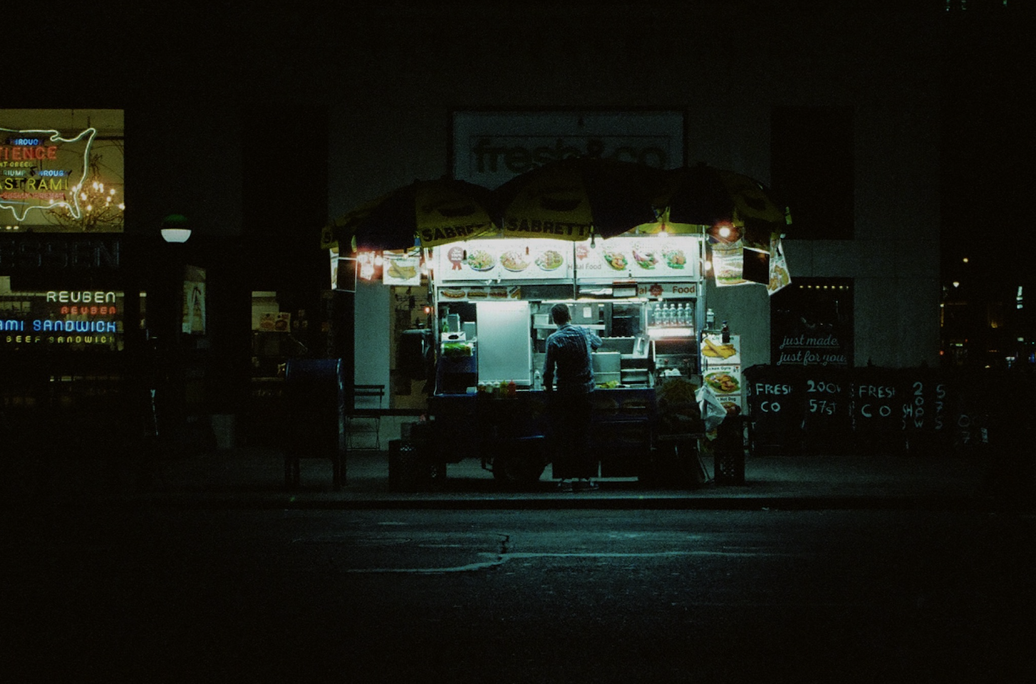 A small food stand illuminated at night, with a person inside preparing food, surrounded by umbrellas and signs displaying menu items.