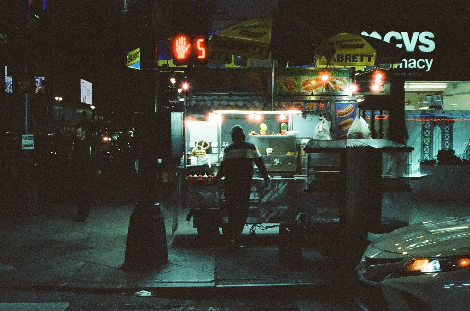 Street food stand at night with a person in front, illuminated by bright lights, with signs displaying hot dog images and promotional banners, and a CVS pharmacy sign in the background.