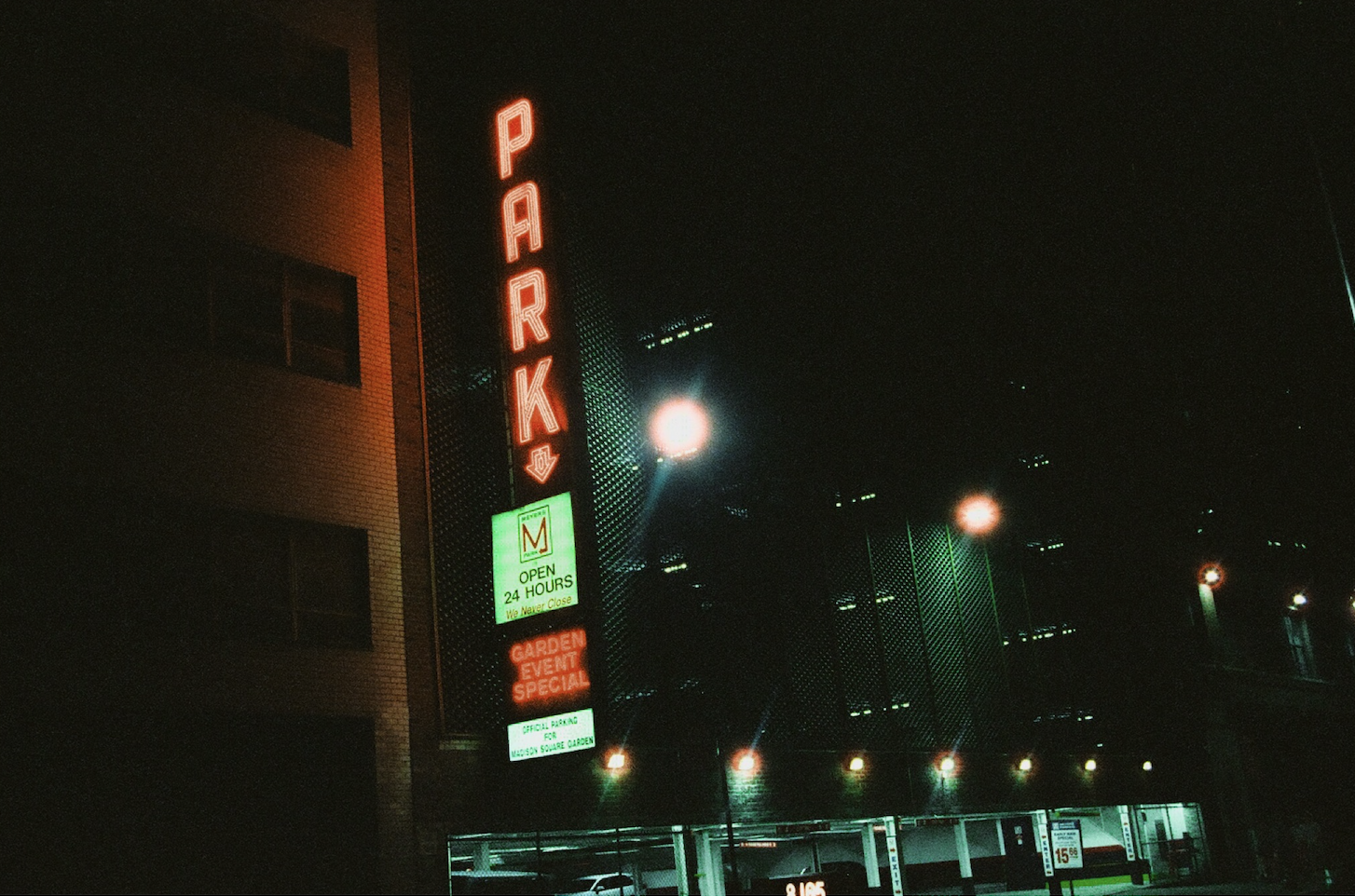 Nighttime street scene with a vertical neon sign reading 'PARK' in red, a lit green and white sign indicating parking open 24 hours, and smaller signs about garden events and official parking.