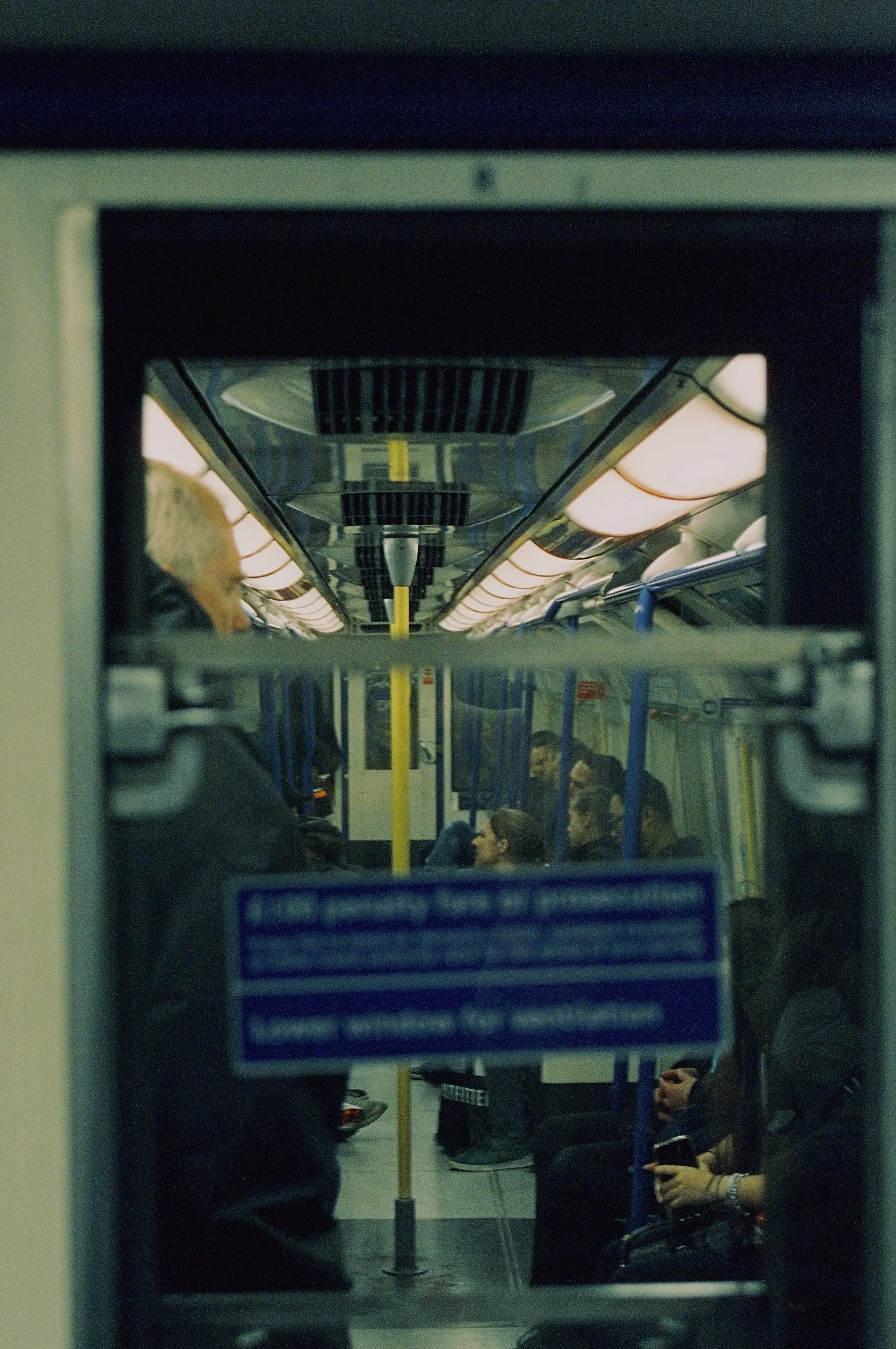 Inside a subway train, view through a window showing passengers seated and standing, with a yellow pole in the center, overhead lighting, and ceiling vents.