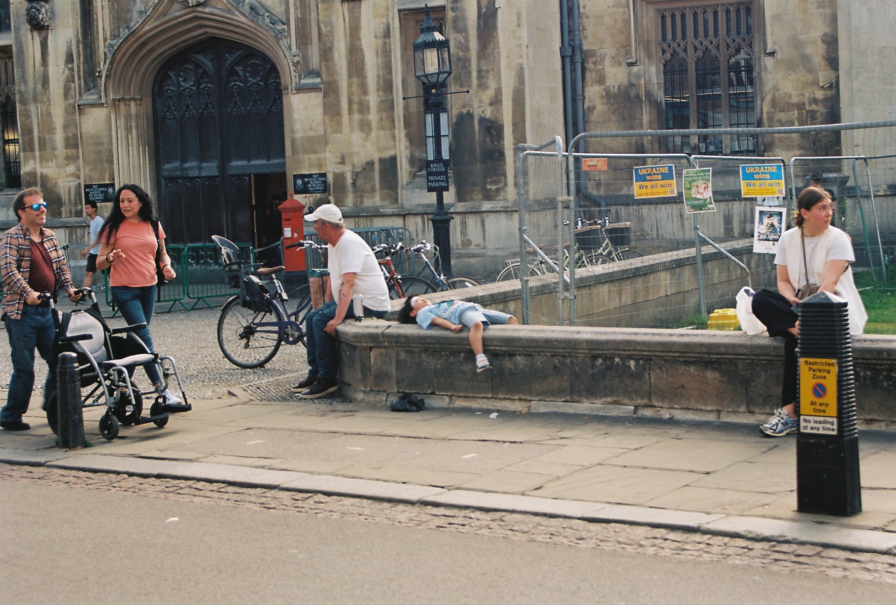 A street scene with several people, including a man sitting on a ledge with a child lying beside him, and a woman sitting alone with a bag nearby. Two women walk past, and bicycles are parked against a railing. There is an old stone building in the background, and signs indicating a parking zone and political messages related to Ukraine.