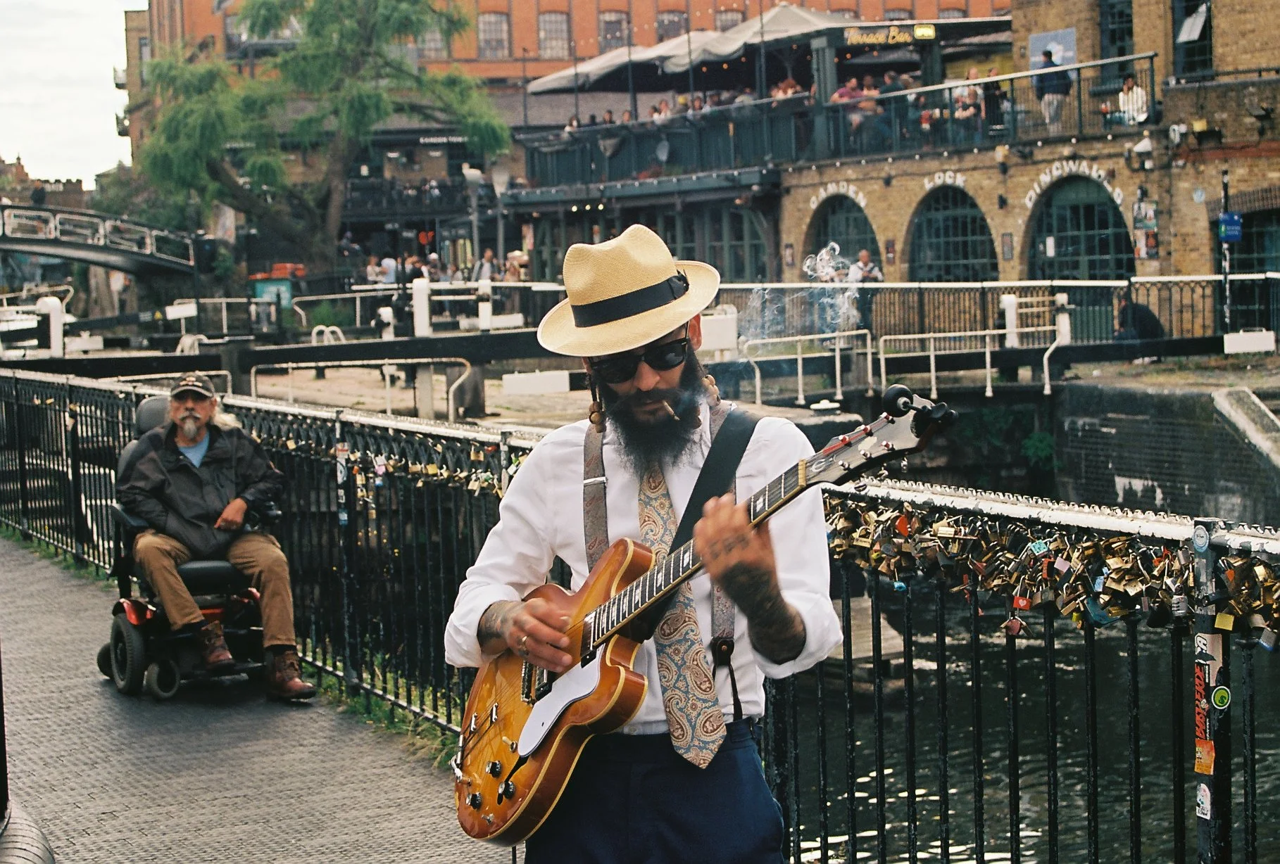 A man wearing a fedora, sunglasses, and a white shirt playing an electric guitar by a canal with a bridge and buildings in the background. A man in a motorized wheelchair is nearby, and there are many locks attached to the bridge's railing.