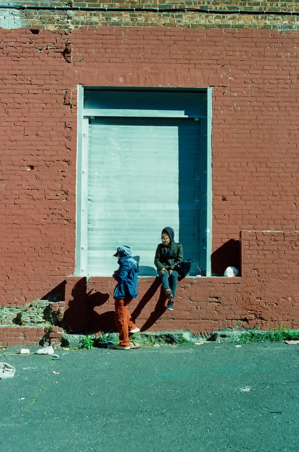 A red brick building with a large metal rolling door, two children in jackets and hats, one sitting on the ledge and the other standing nearby, engaged in conversation, with shadows cast on the wall.