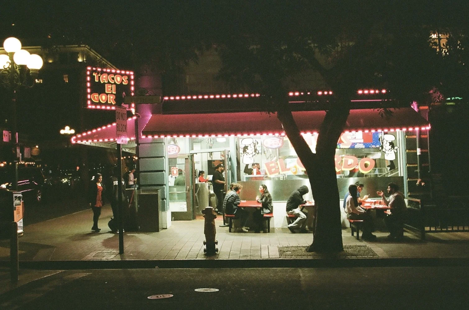 Nighttime scene at a taco stand called 'Tacos El Gordo' with illuminated red neon signs, several customers sitting at red tables, and a few employees working inside, with a tree and streetlamp visible in the foreground.