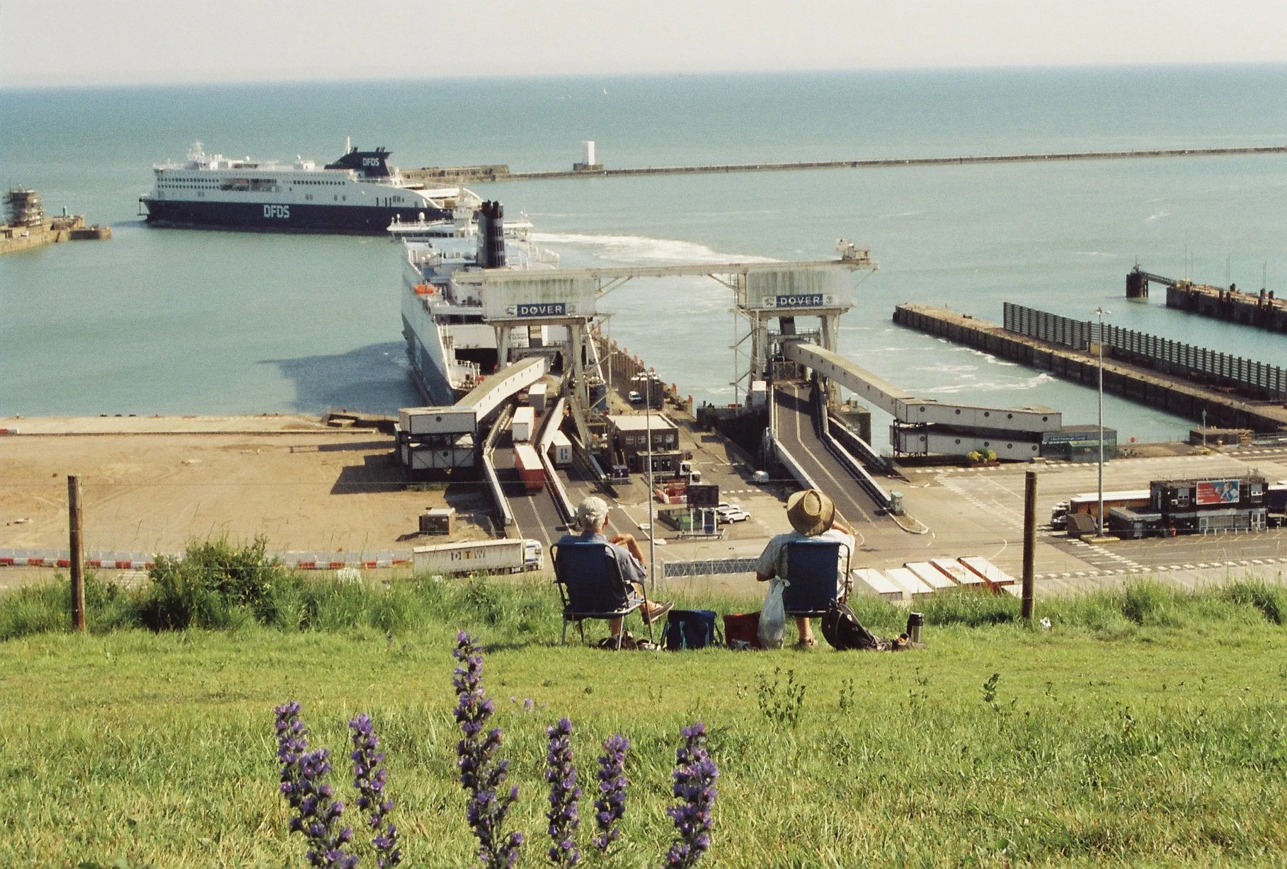 Two people sitting on chairs on a grassy hill overlooking a harbor with ferry docks and ships in Dover.