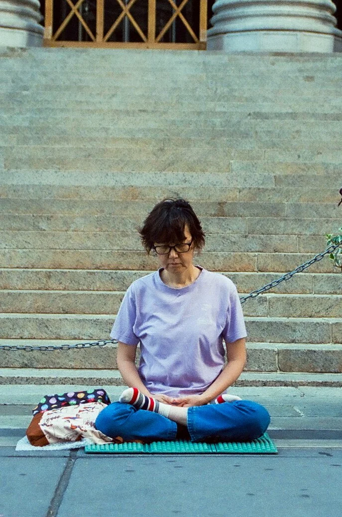 A woman sitting cross-legged on a mat on the sidewalk, with her eyes closed and meditating, next to a backpack and a t-shirt, in front of stone stairs and a chain barrier.