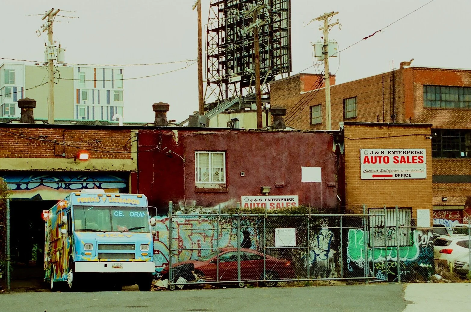 A red brick building with an 'Auto Sales' sign, a colorful truck parked in front, surrounded by graffiti fencing, and power lines overhead in an urban area.
