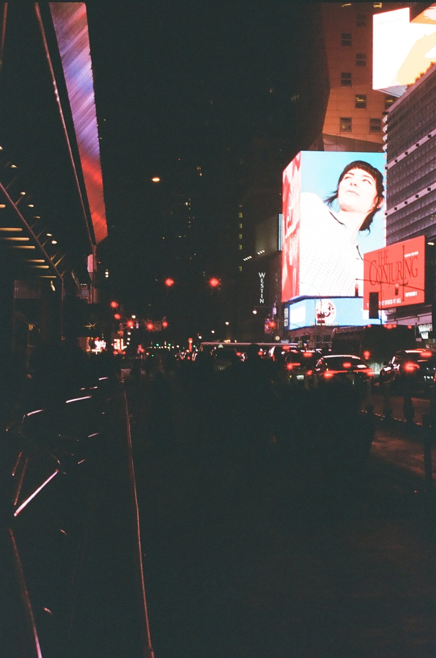 Nighttime city street scene with bright electronic billboards and lots of cars, in an urban area. One large digital billboard displays a person's face.