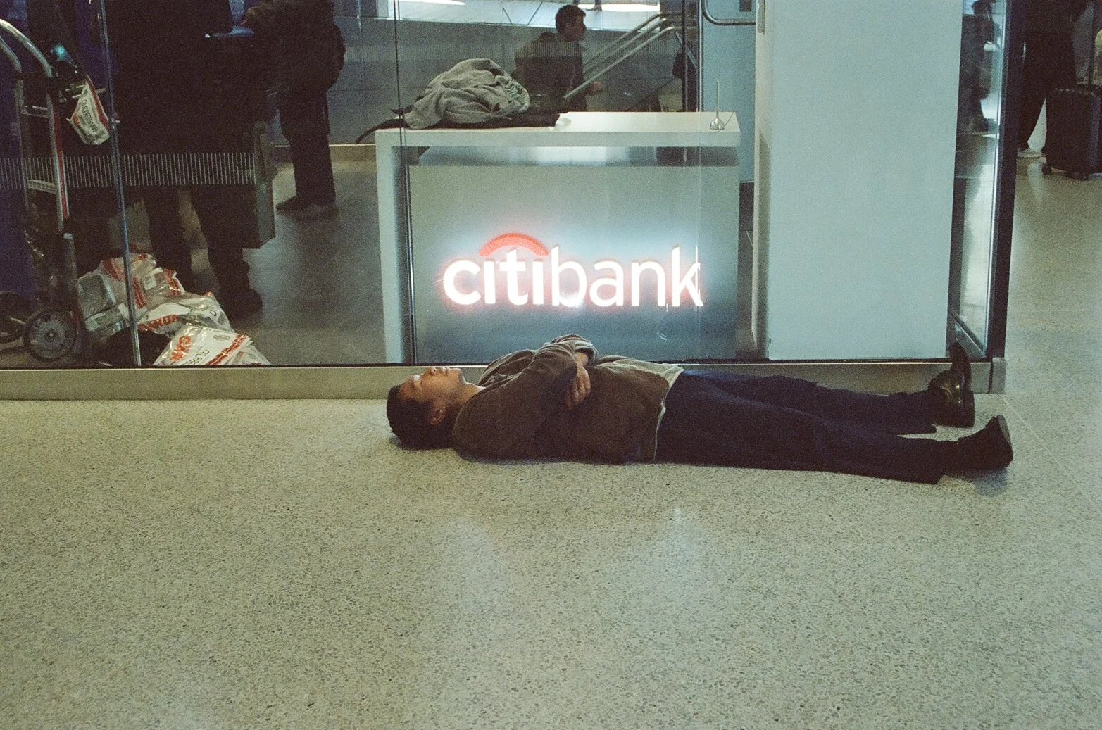 A man lying on the floor of an airport near a glass wall with a lit Citibank sign behind him.