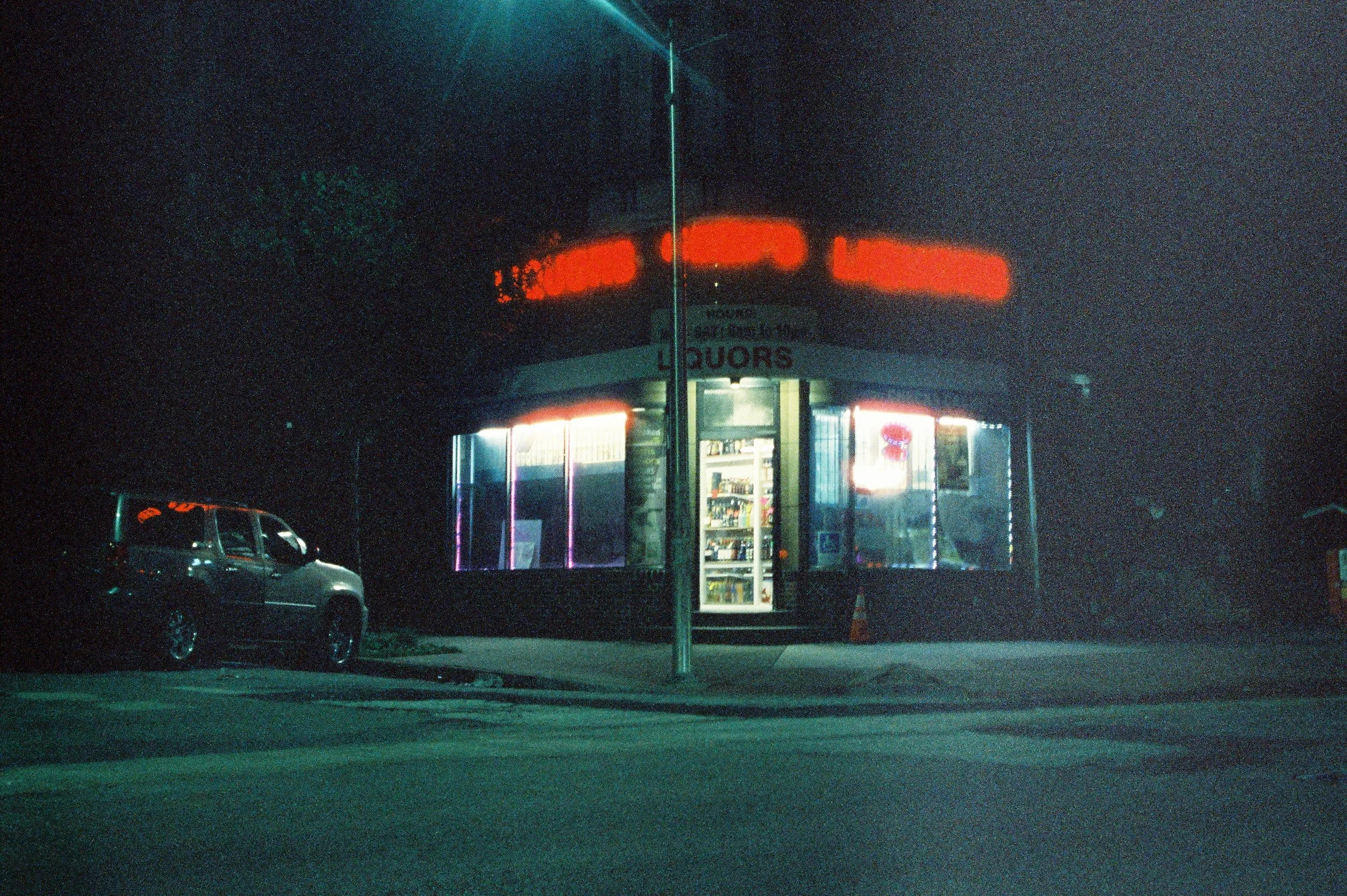 Nighttime view of a corner convenience store with bright neon lights, a visible cash register inside, and a parked car in front.