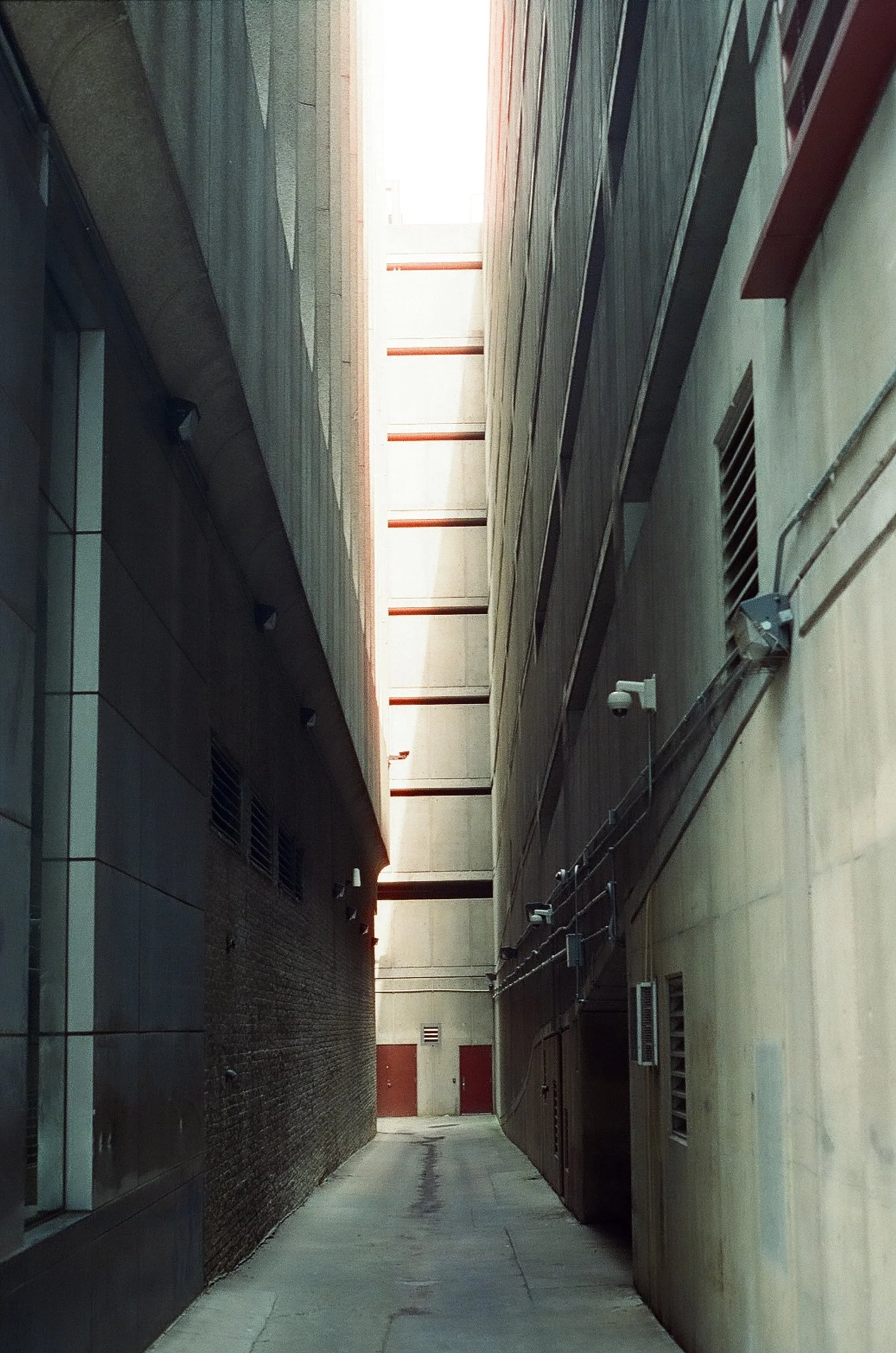 Narrow alleyway between tall concrete and brick buildings, with exposed pipes and security cameras, leading to a small area with red doors and bright light at the end.