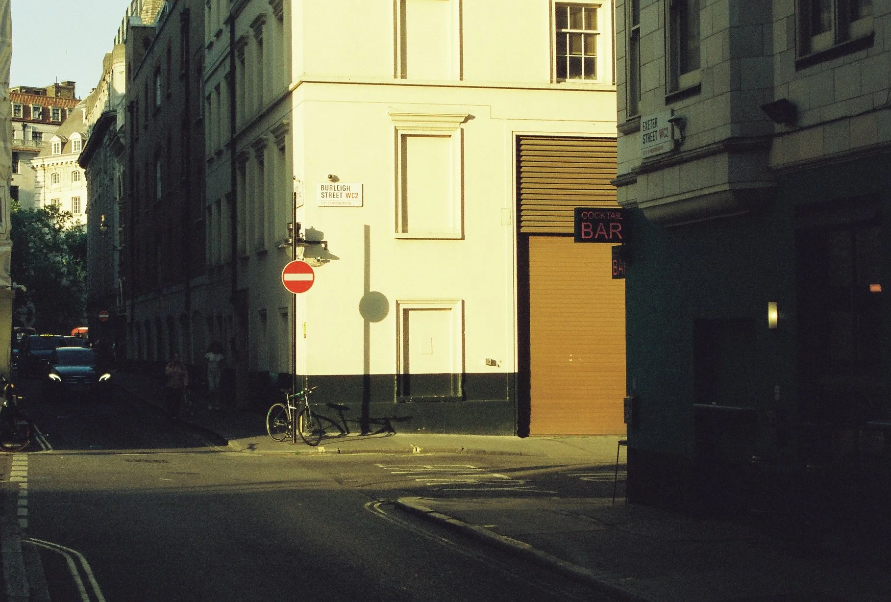 A street corner with a 'No Entry' sign, bicycles parked against a lamp post, and a building with a sign reading 'Cocktail Bar.' The street signs indicate 'Burleigh Street WC2' and 'Exeter Street WC2.' The scene is in sunlight, casting shadows on the buildings and street.